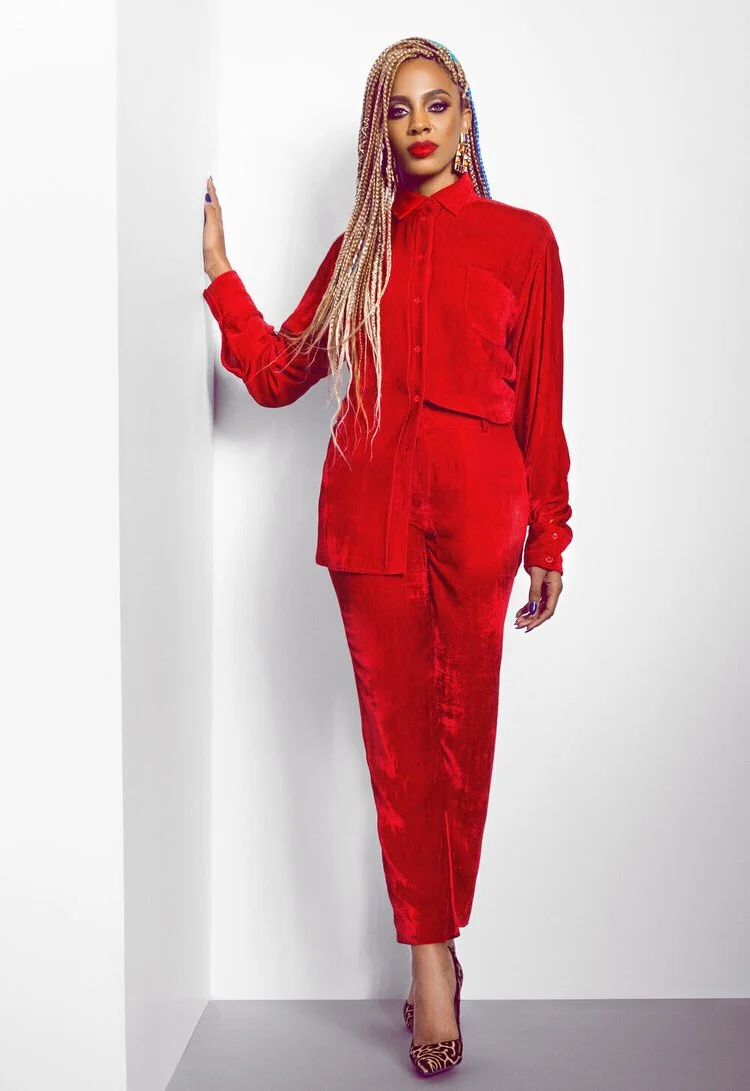 Woman with braided multicolor hair wearing a red velvet shirt and pants, red lipstick, and leopard print high heels, posing against a white wall.