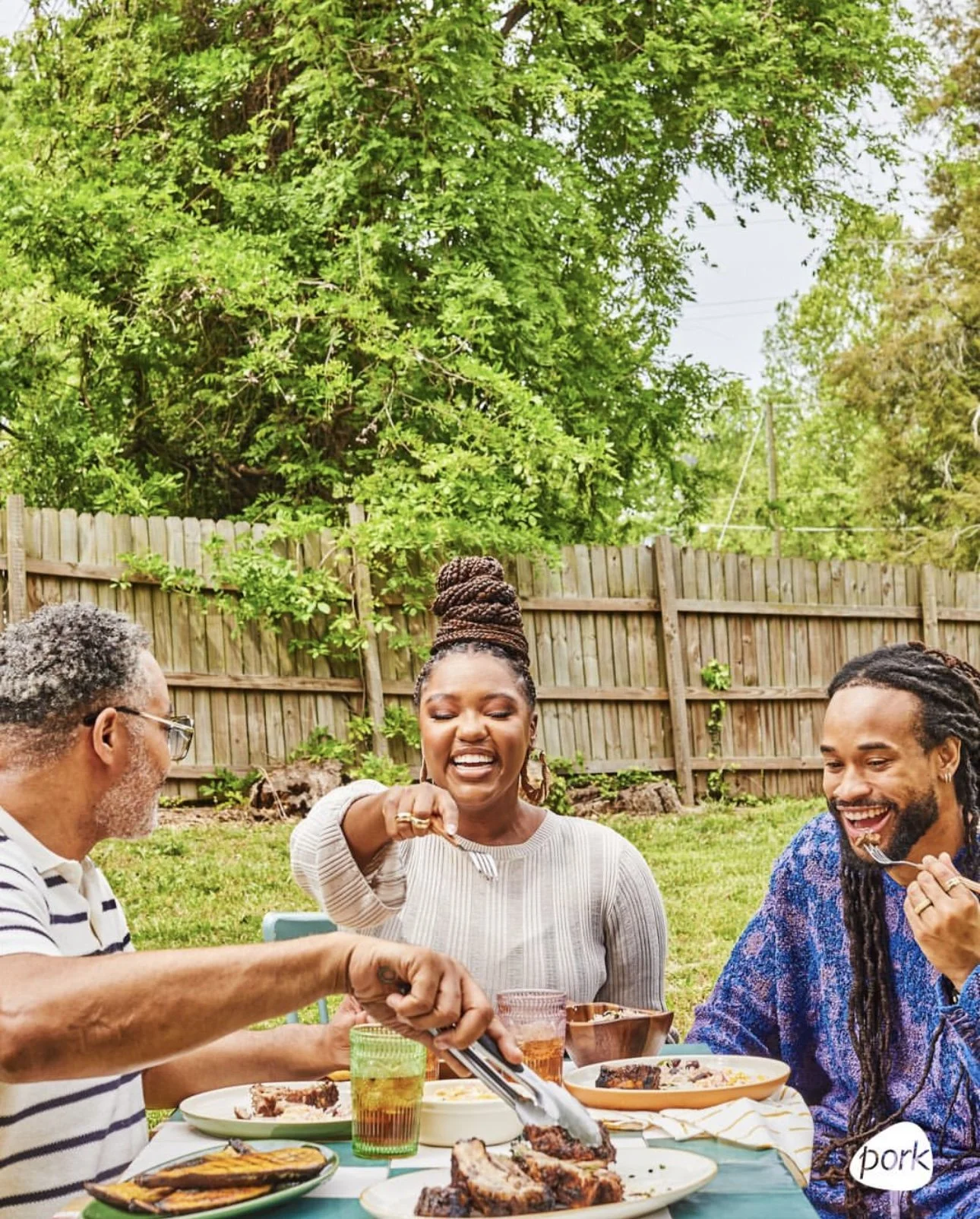 Three people enjoying a backyard barbecue, with one person serving food and the other two smiling and eating, on a sunny day with trees and a wooden fence in the background.