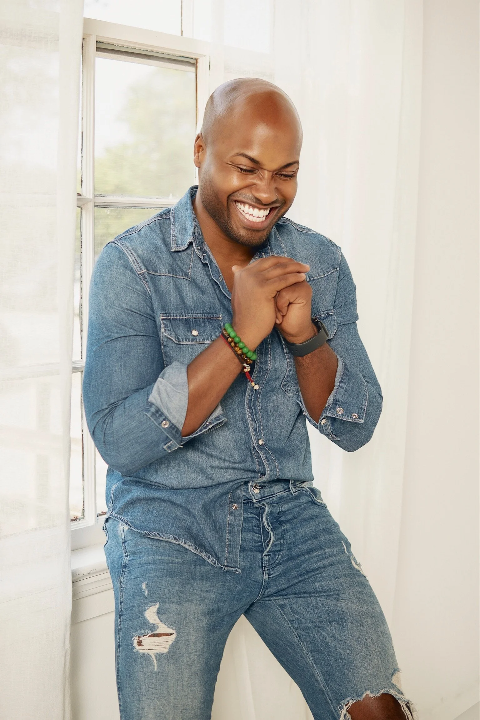 A smiling man in a denim shirt and ripped jeans standing indoors next to a window with white curtains.