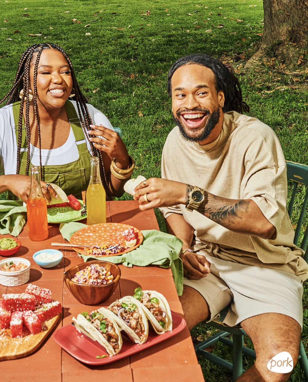 Two people enjoying an outdoor meal at a picnic table, with various dishes including tacos, a salad, and drinks, on a grassy area with a tree in the background.