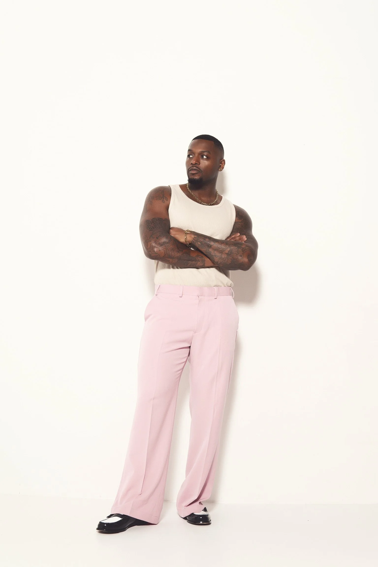 A man with tattoos on his arms wearing a white sleeveless shirt, pink wide-leg trousers, and black-and-white shoes, standing against a white wall with arms crossed.