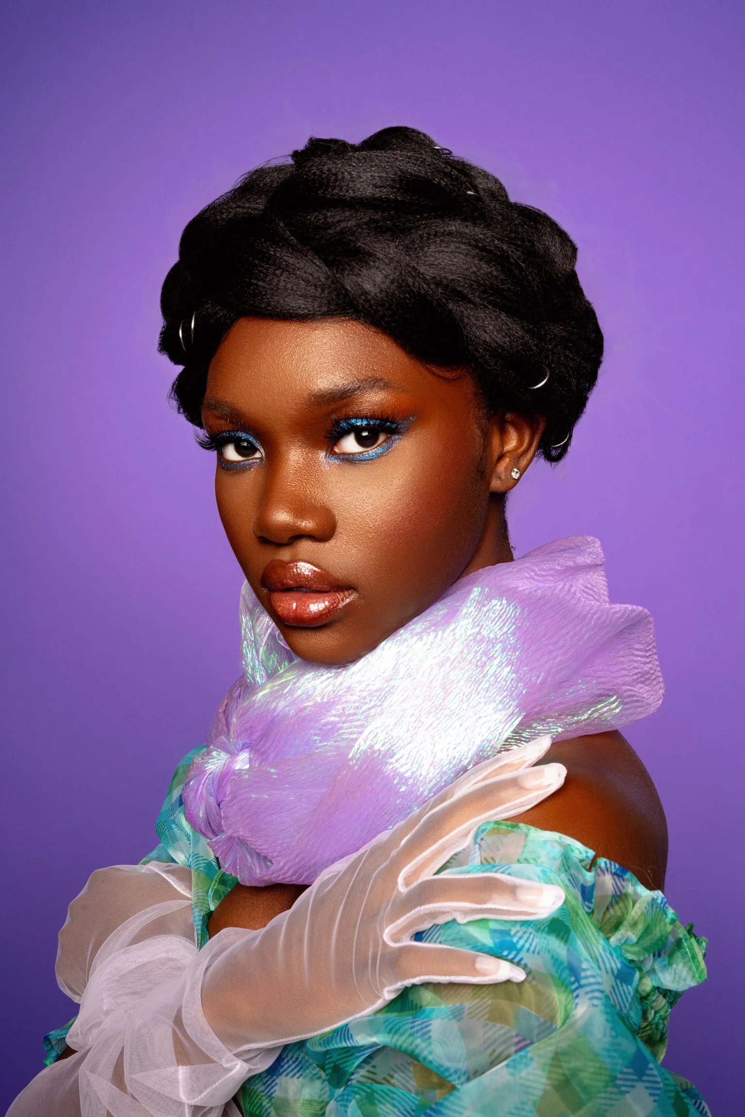A young woman with dark skin and short black hair styled with hair clips, wearing bold blue eye makeup and glossy lips, posed against a purple background, wearing iridescent and patterned clothing with sheer gloves.