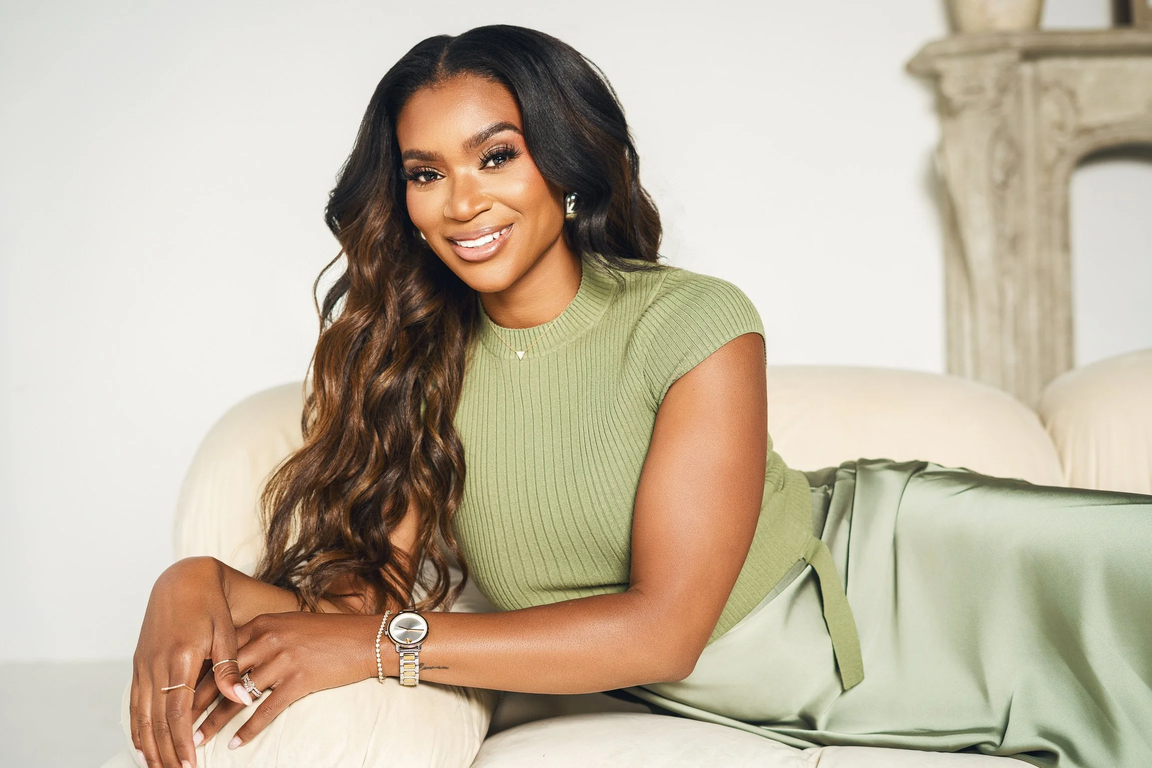 A woman with long wavy hair, wearing a green sleeveless top and a matching green skirt, lying on a cream-colored couch, smiling at the camera.