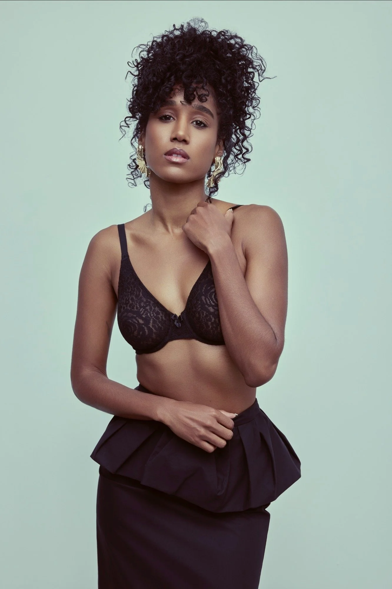 A woman with curly hair wearing gold earrings, a black lace bra, and a black skirt, posing against a plain light green background.