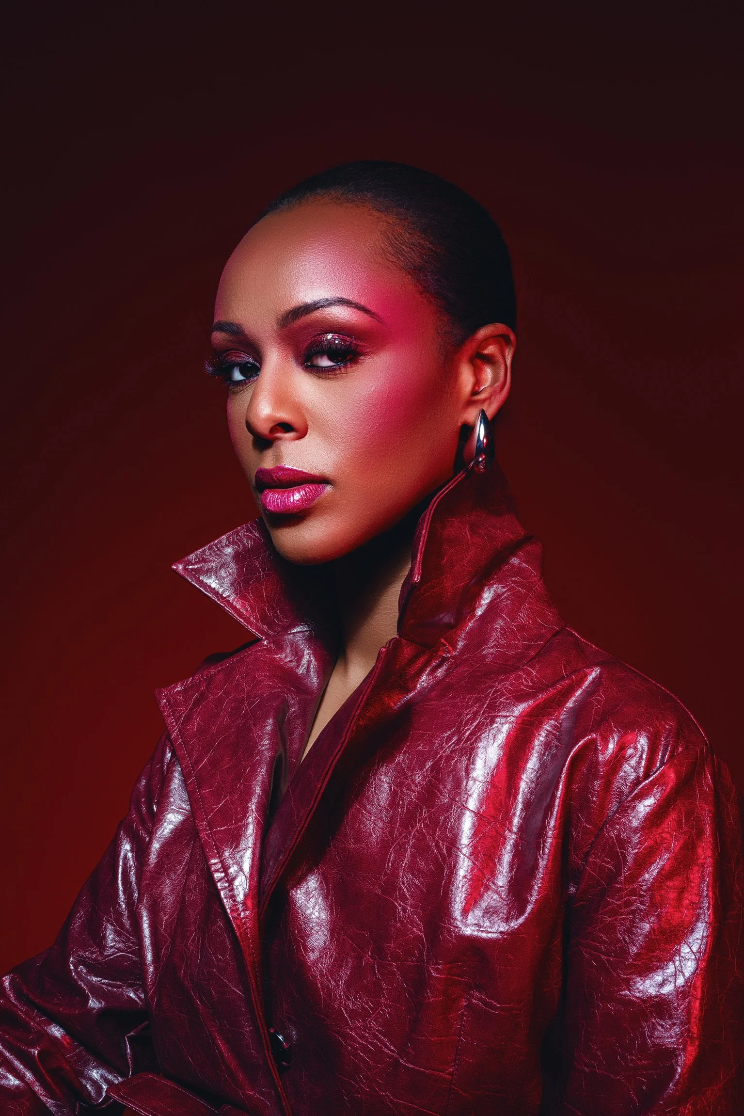 A woman with a bold makeup look and short hair wearing a shiny red leather jacket against a dark red background.