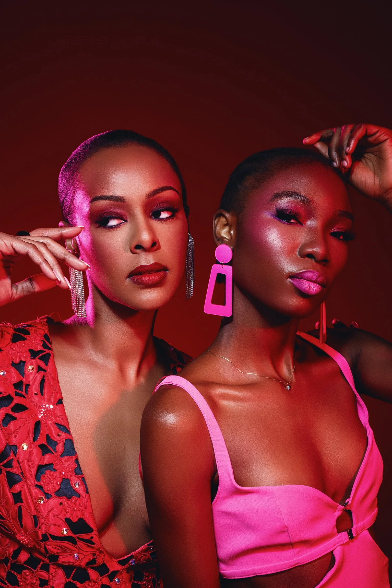 Two women with short hair and colorful makeup posing against a red background, wearing vibrant outfits and statement earrings.