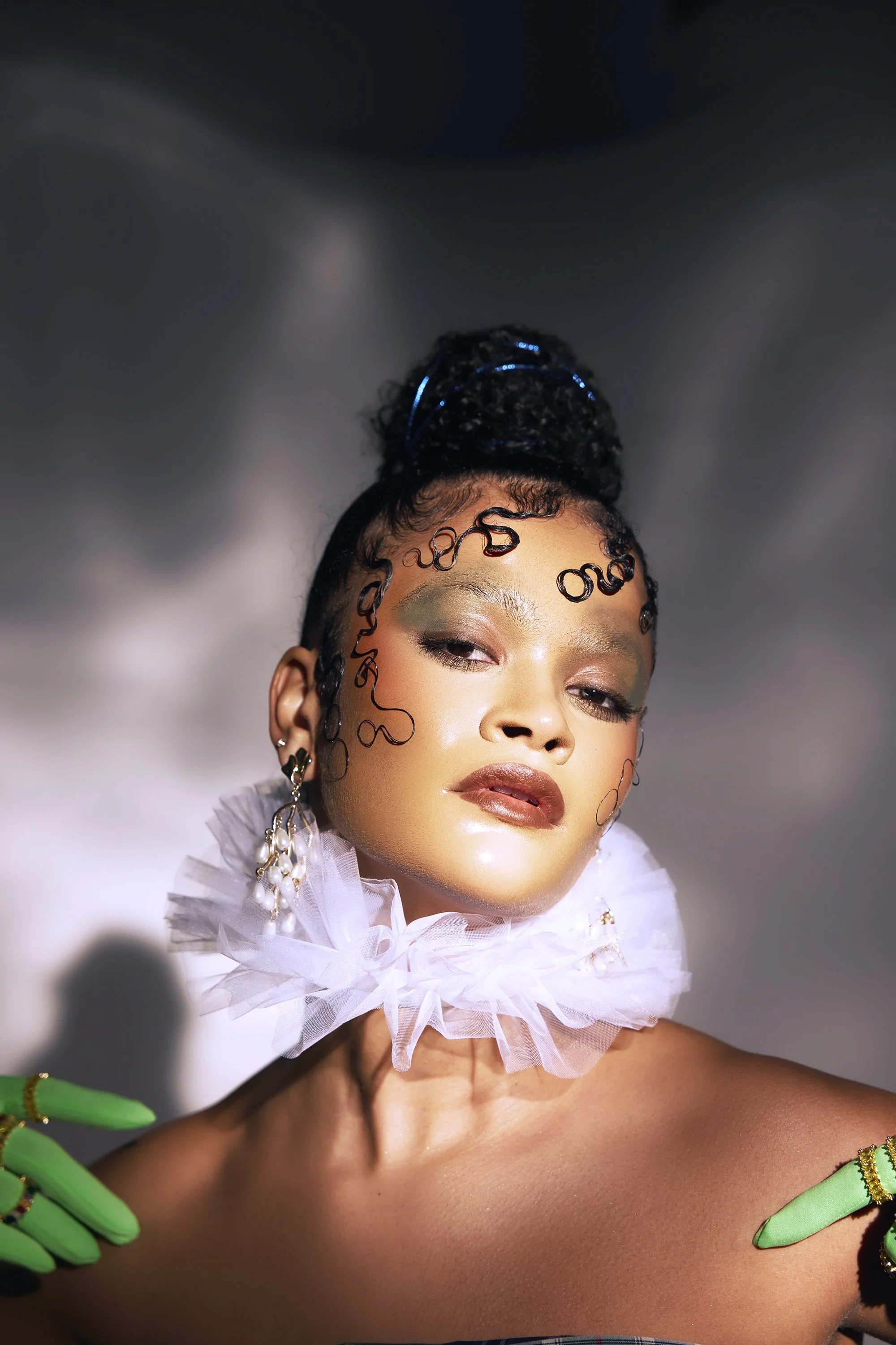 Portrait of a woman with curly hair styled in an updo, wearing dramatic makeup, a white ruffled collar, green gloves with gold accents, and large pearl earrings, against a blurred dark background.