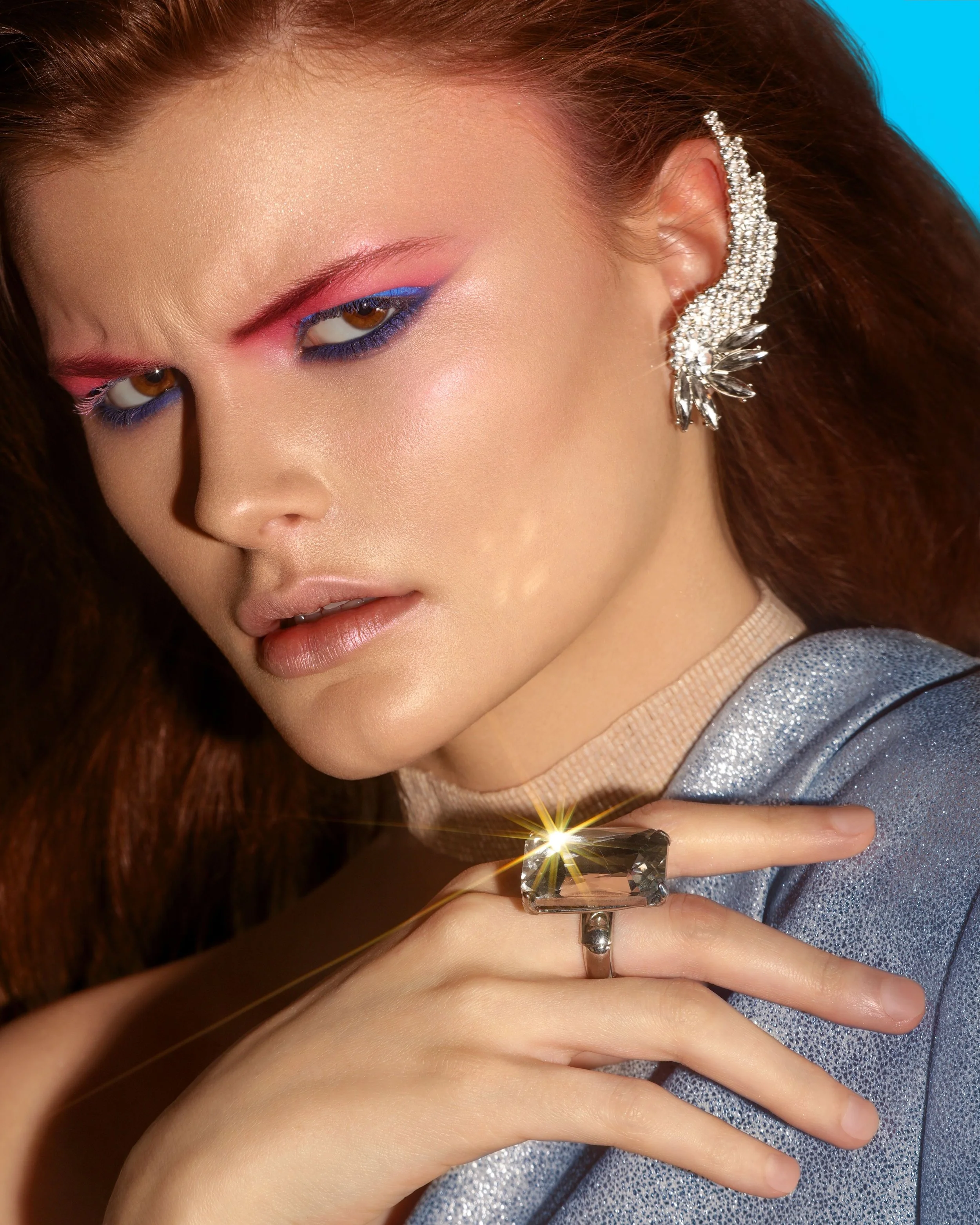 Close-up of a woman with light makeup, colorful eyeshadow, wearing a sparkly silver jacket, a large gemstone ring, and an elaborate pearl and crystal earring, with a bright blue background.