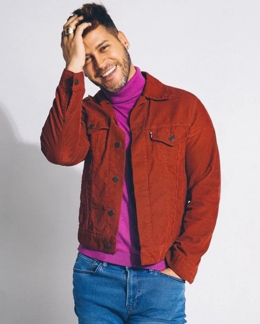 A man with dark hair and a beard, smiling and touching his head, is wearing a purple turtleneck, a red denim jacket, and blue jeans.
