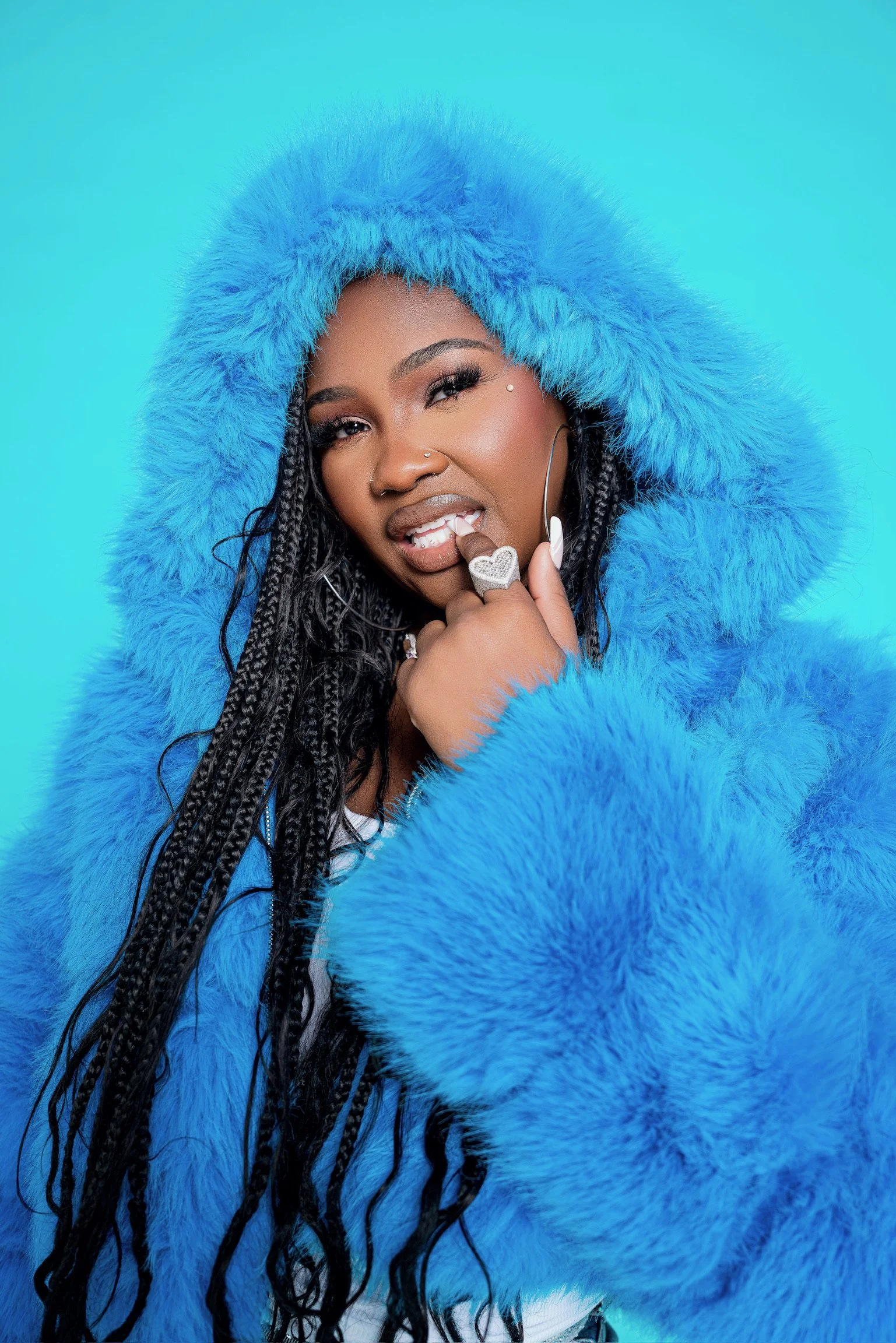 A woman with long braided hair wearing a bright blue faux fur hood and coat, posing with a playful expression against a blue background.