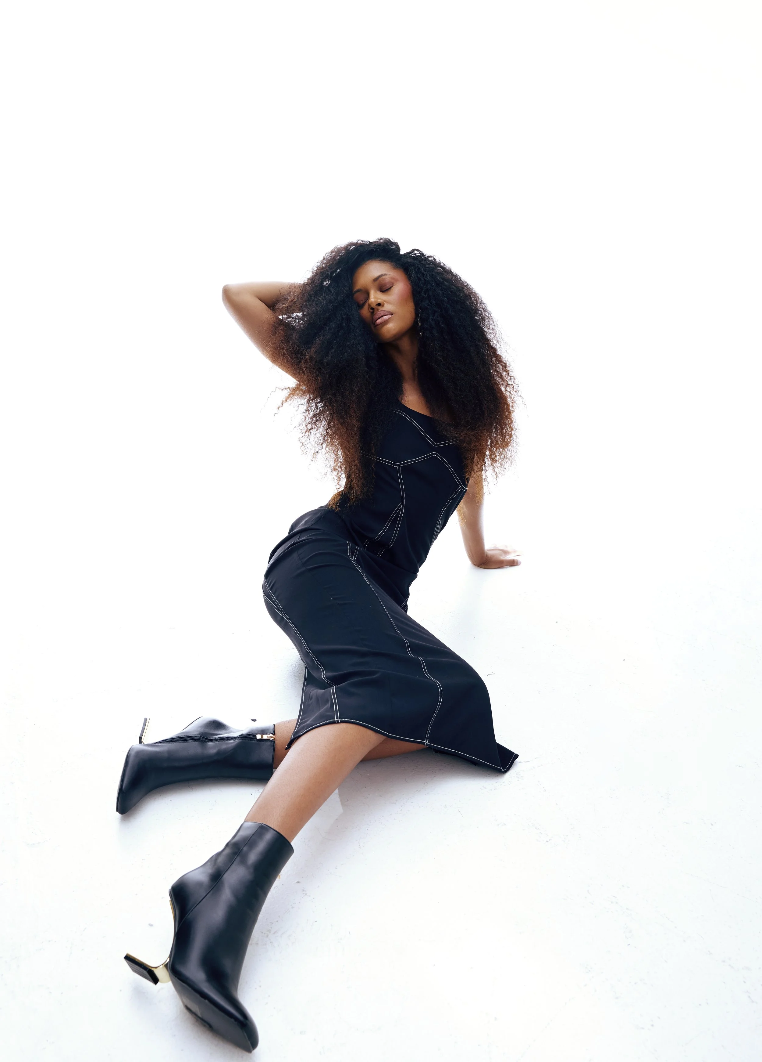 Woman with long curly hair wearing a black dress and black ankle boots, sitting on the floor with one arm behind her and her eyes closed.