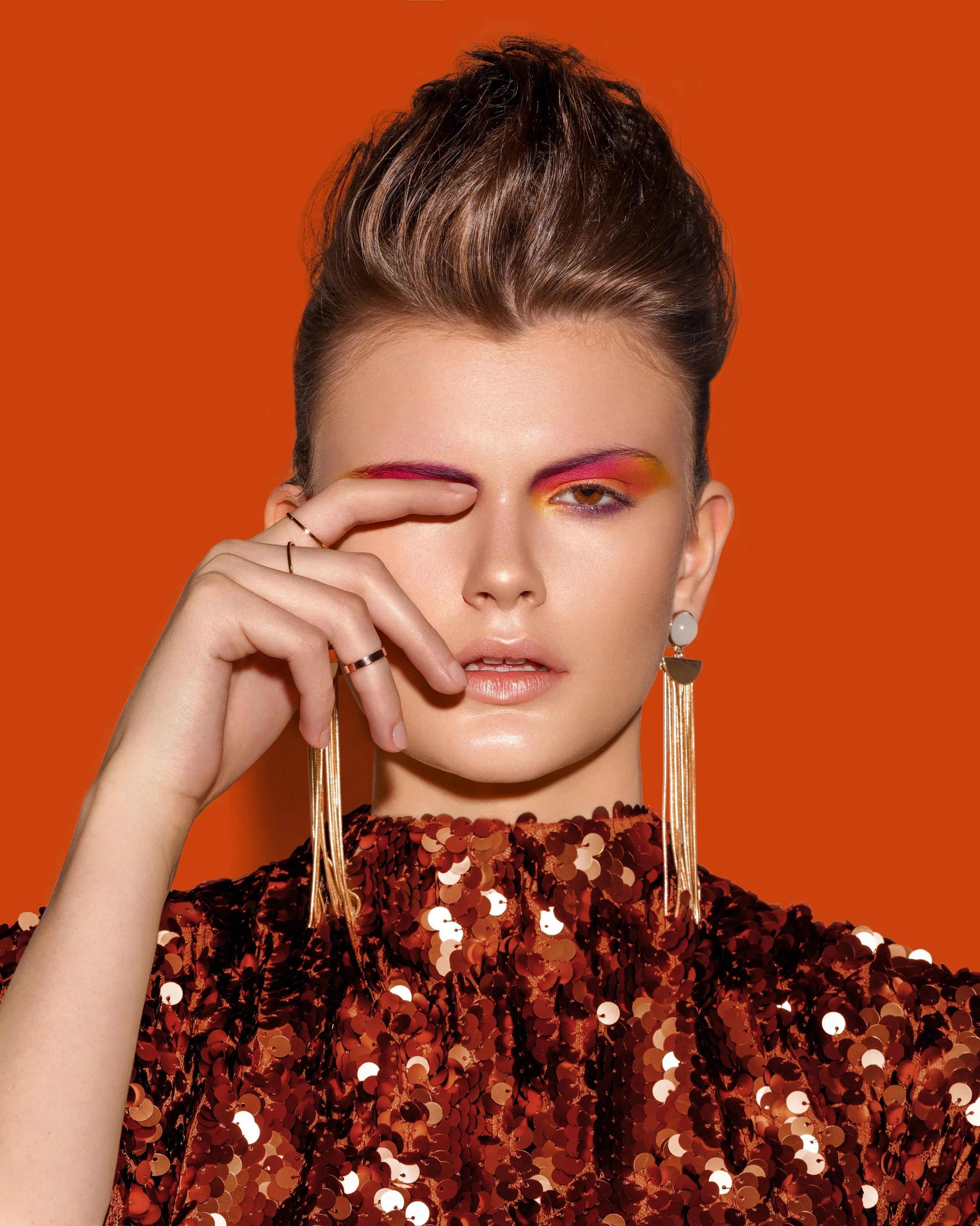 A woman with short brown hair styled up, wearing bright makeup with red and yellow eyeshadow, and a shiny sequined red top. She is holding her hand near her face, with rings on her fingers, and long dangling earrings. The background is solid orange.