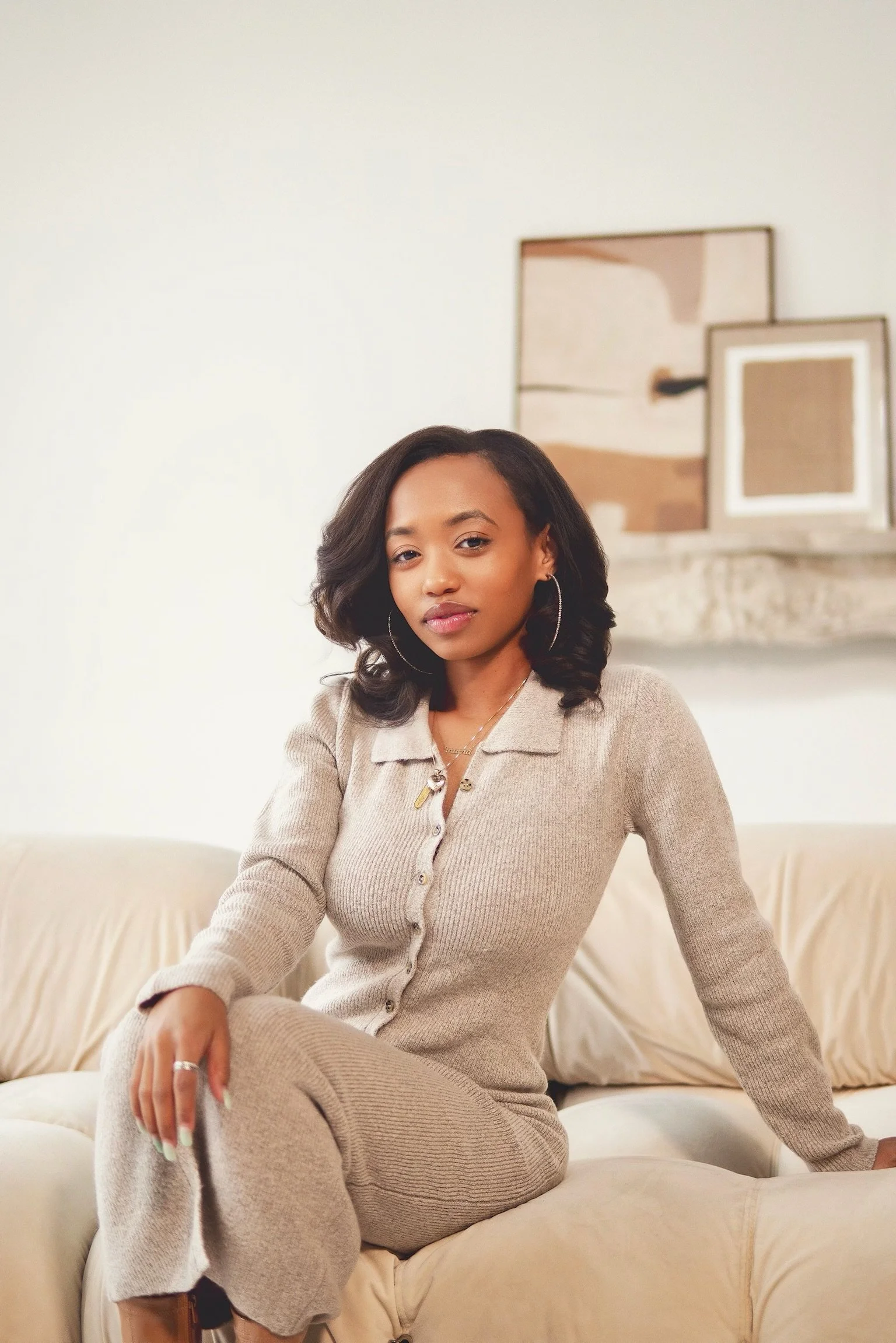 A woman wearing a beige knit cardigan and pants sitting on a cream-colored sofa, with a neutral expression and stylish hair, in a living room with abstract artwork on the wall behind her.