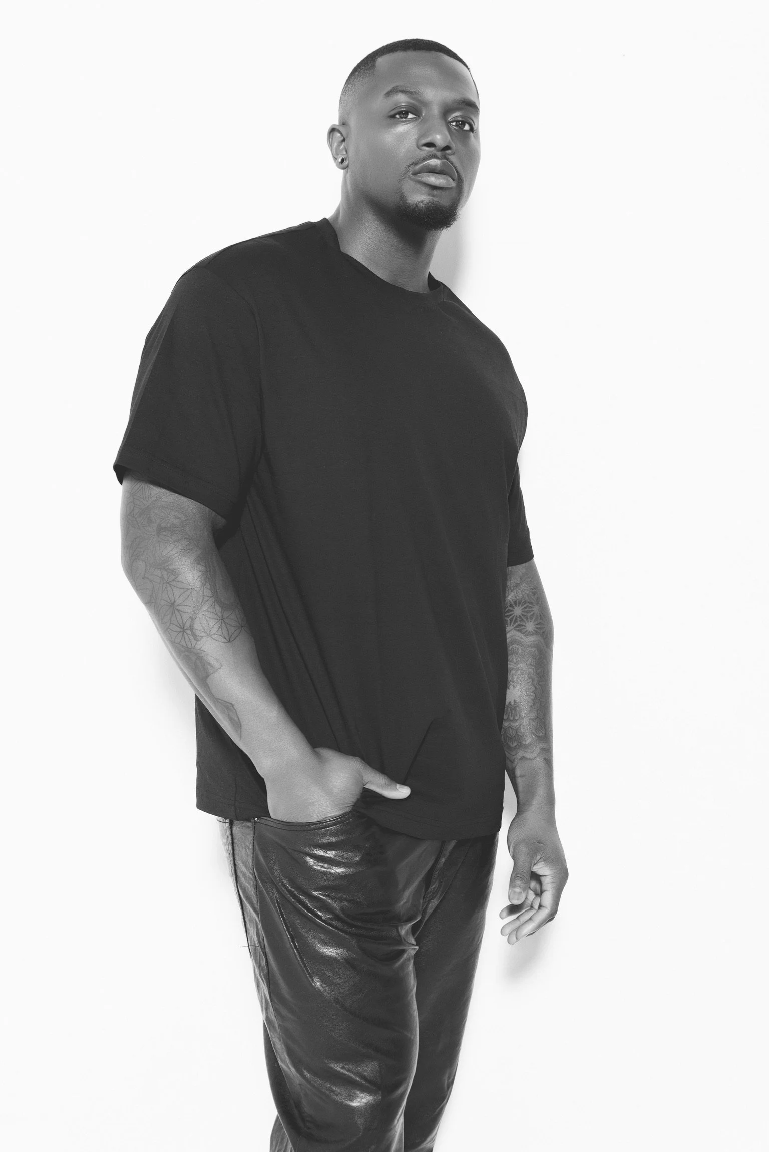 A man with tattoos on his arms, wearing a black t-shirt and shiny dark pants, standing against a plain white background.