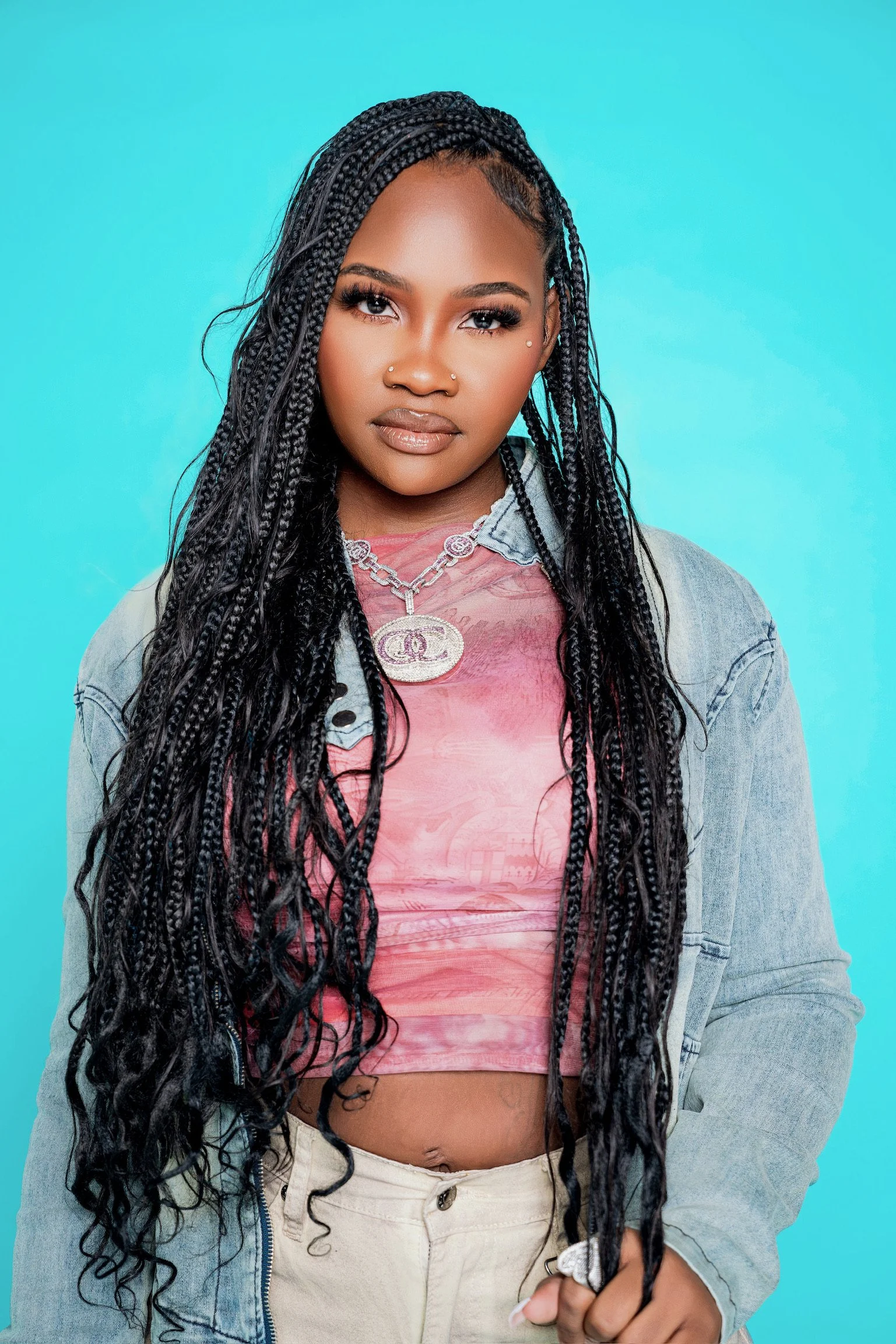 Young woman with long braids, wearing a pink crop top, necklace, and light denim jacket, standing against a blue background.