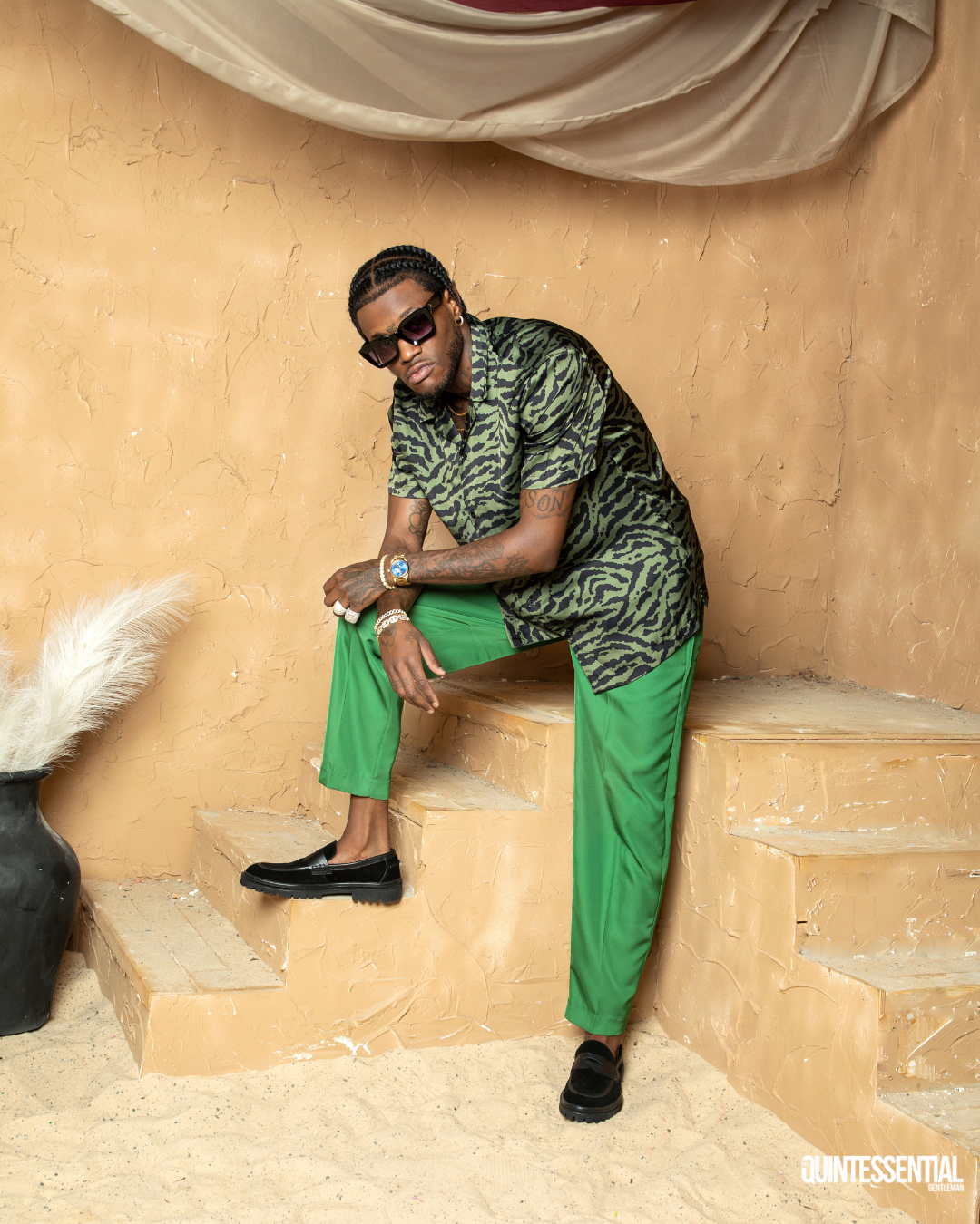 A man with braided hair wearing sunglasses, a tiger-striped shirt, bright green pants, and black loafers, sitting on beige steps in a studio with a textured beige wall and a large vase with pampas grass.