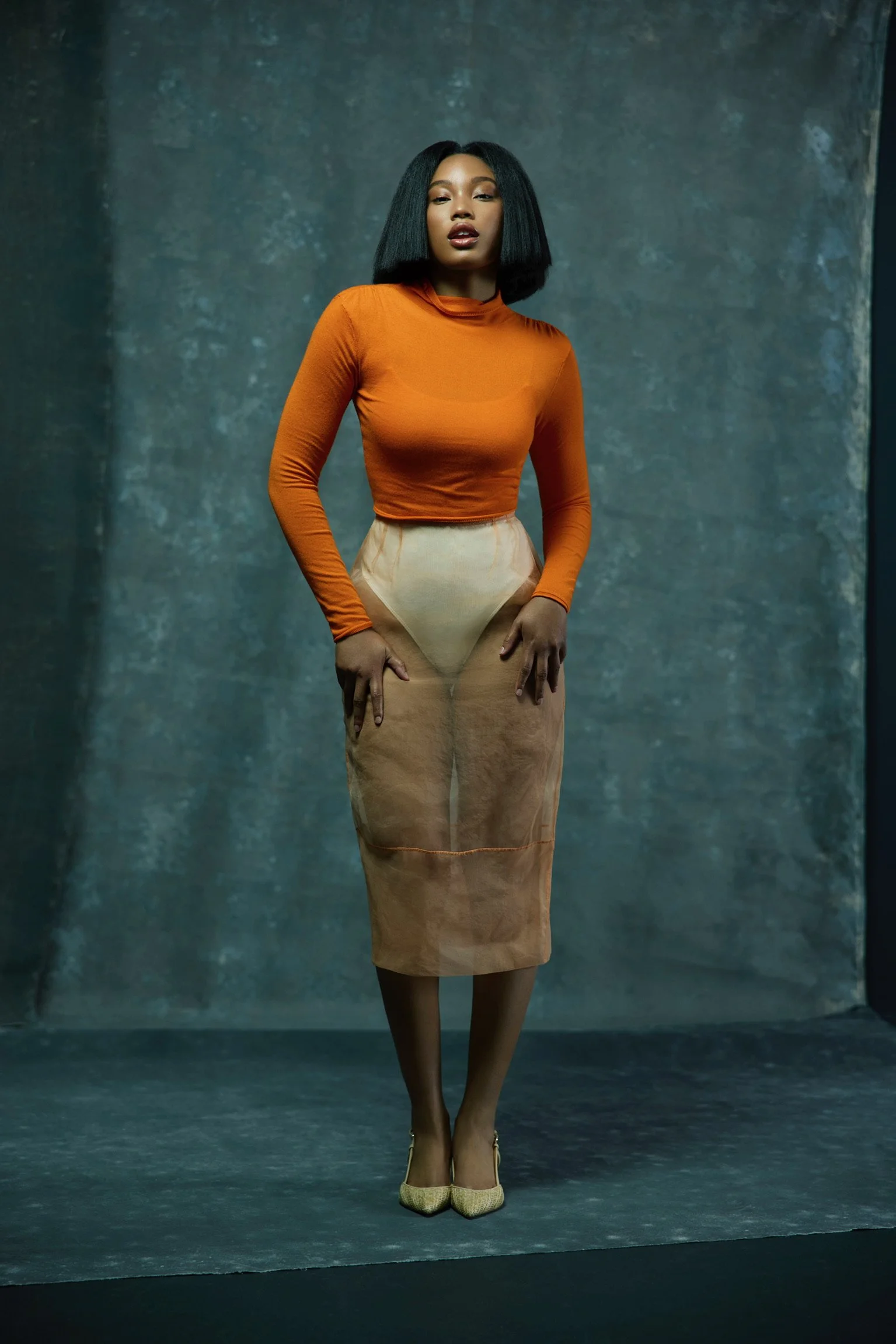 A woman with shoulder-length black hair wearing an orange long-sleeve top, beige sheer skirt, and high heels, standing against a textured dark background.