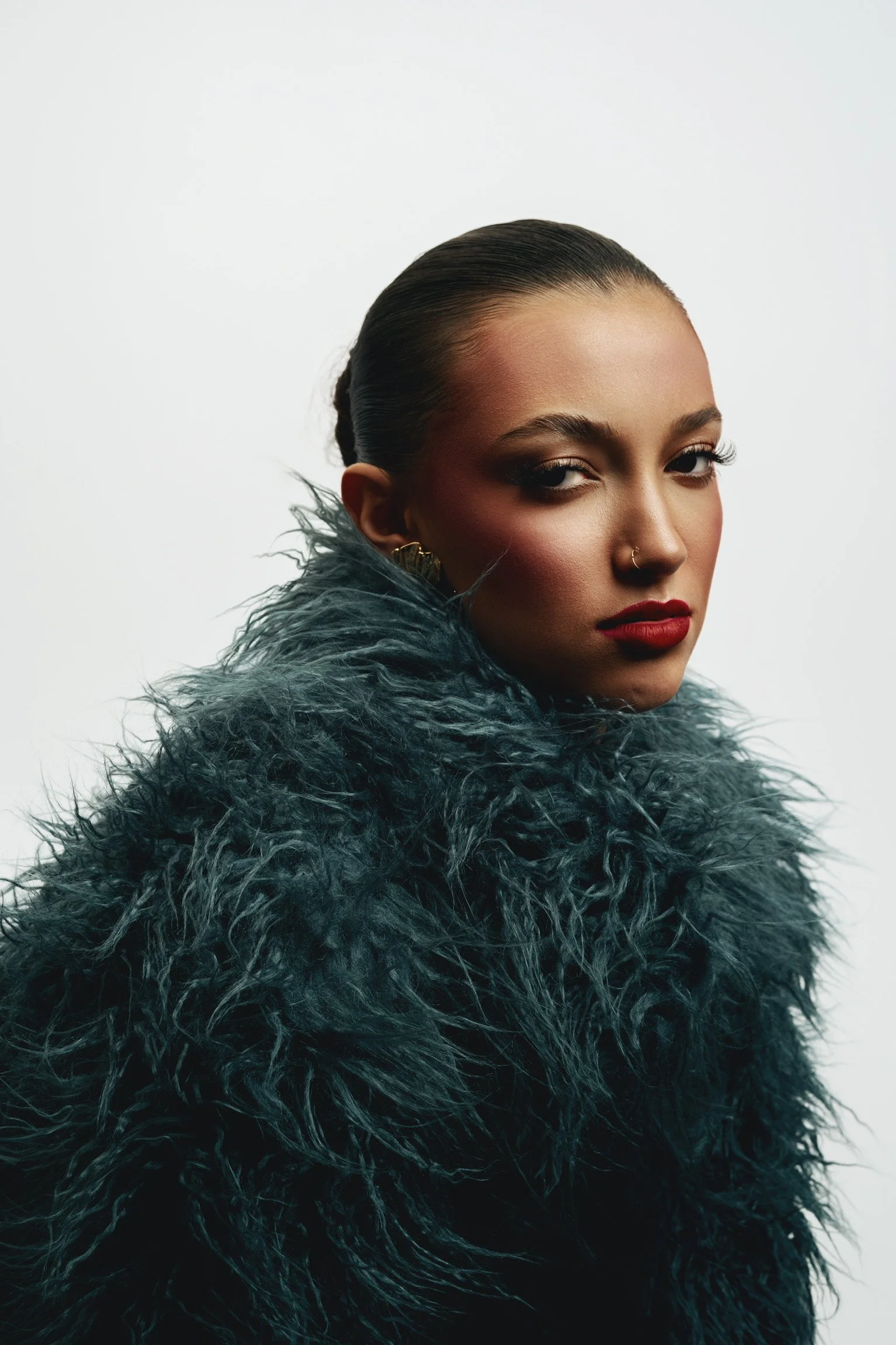 A woman with slicked-back hair wearing a dark, fluffy fur coat looking at the camera against a plain white background.