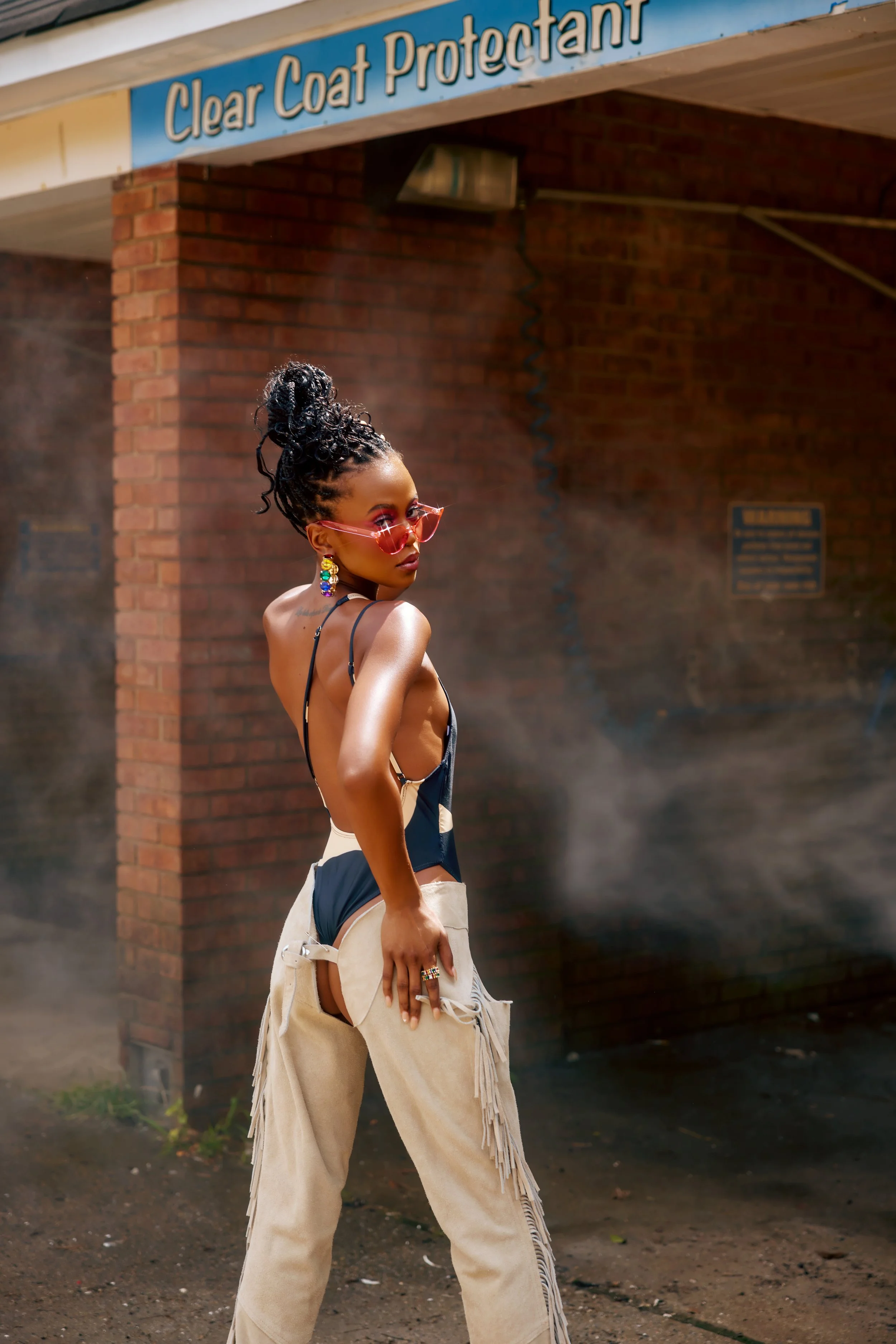 A woman with dark skin and braided hair styled in a high bun, wearing pink sunglasses, colorful earrings, and a revealing black and beige outfit, stands confidently outside a brick building at a car wash facility named 'Clear Coat Protectant'.