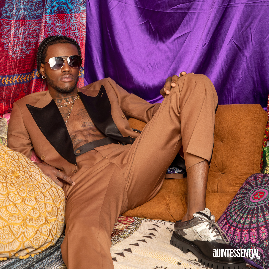 Man reclining on a sofa in colorful, patterned cushions and fabrics, wearing sunglasses, a tan suit with a black lapel, and chunky black and beige shoes, with a purple backdrop.