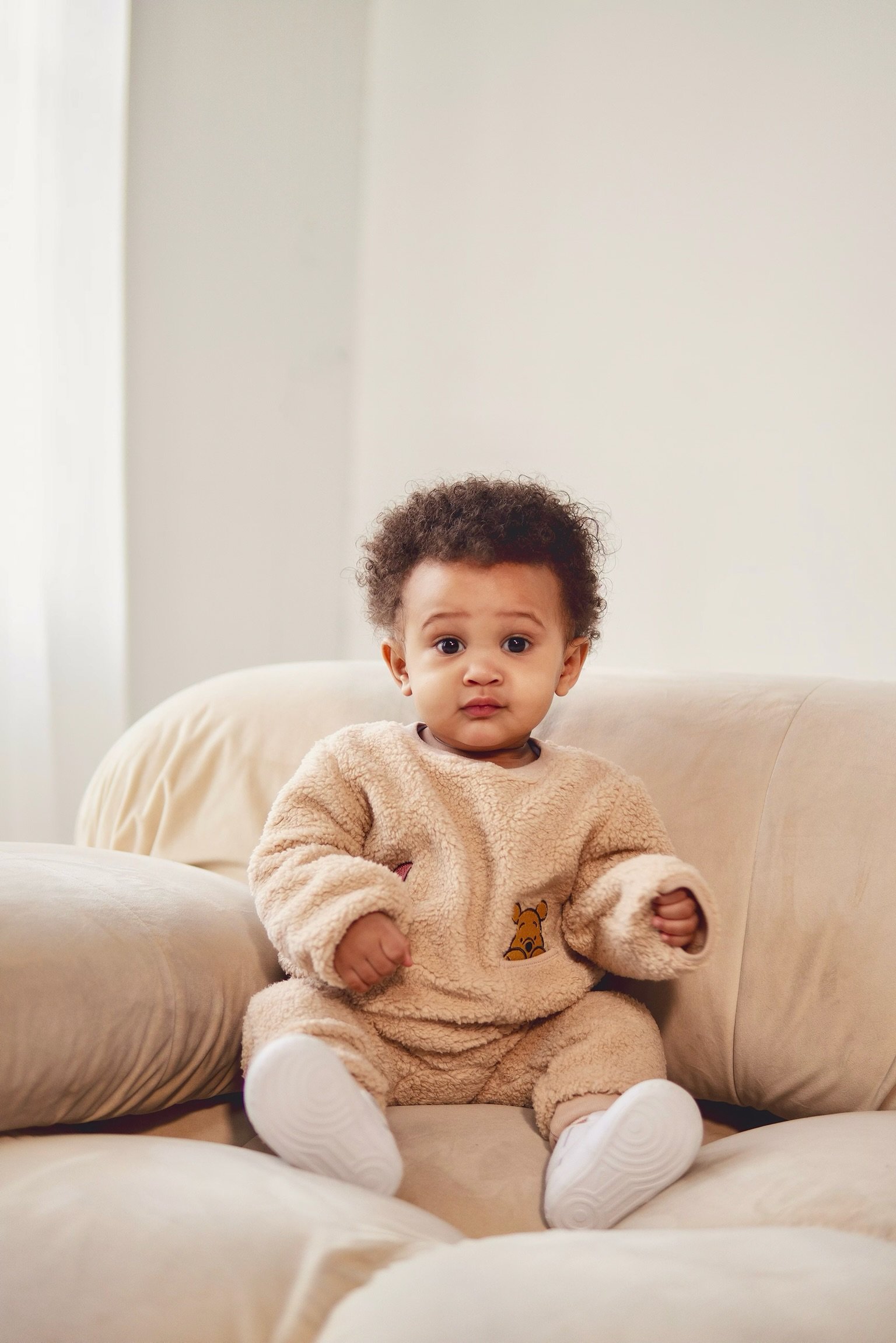 A young child with curly hair sitting on a beige couch, wearing a plush beige outfit with a giraffe print, and white shoes, looking at the camera with a curious expression.