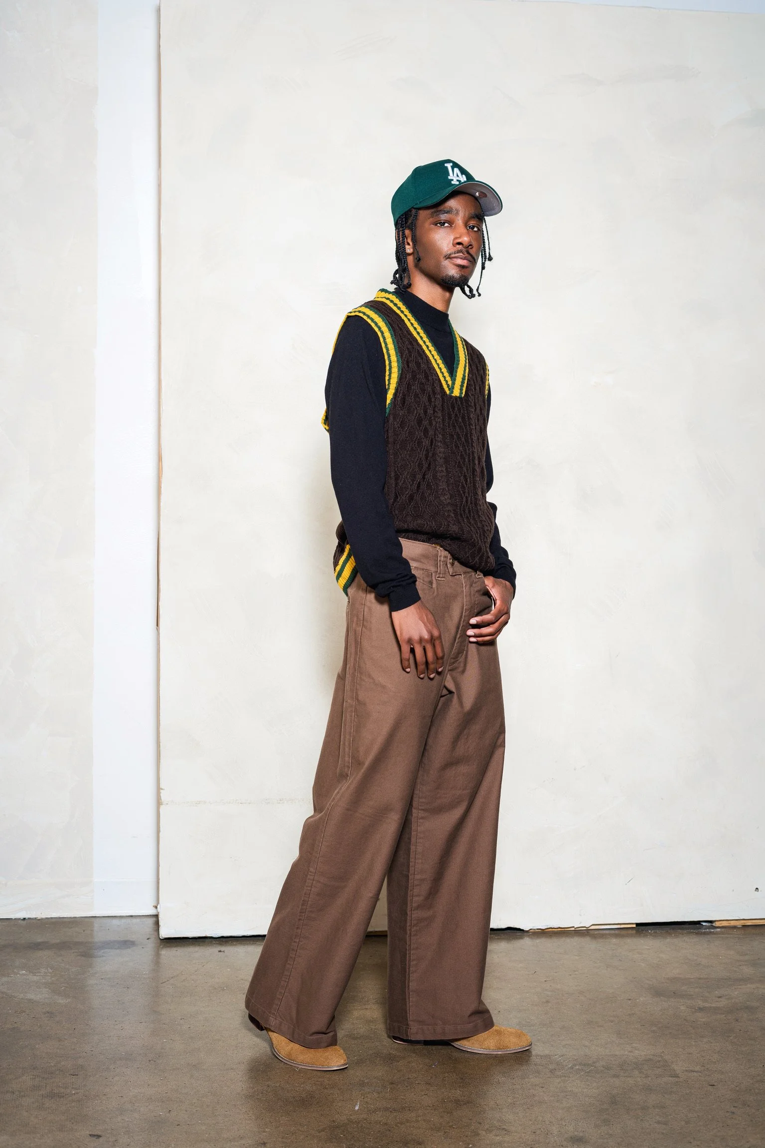 Young man standing indoors against a plain light-colored wall, wearing a gray baseball cap, black long-sleeve shirt, brown wide-leg pants, beige shoes, and a dark sleeveless sweater with yellow and green stripes on the collar and armholes.