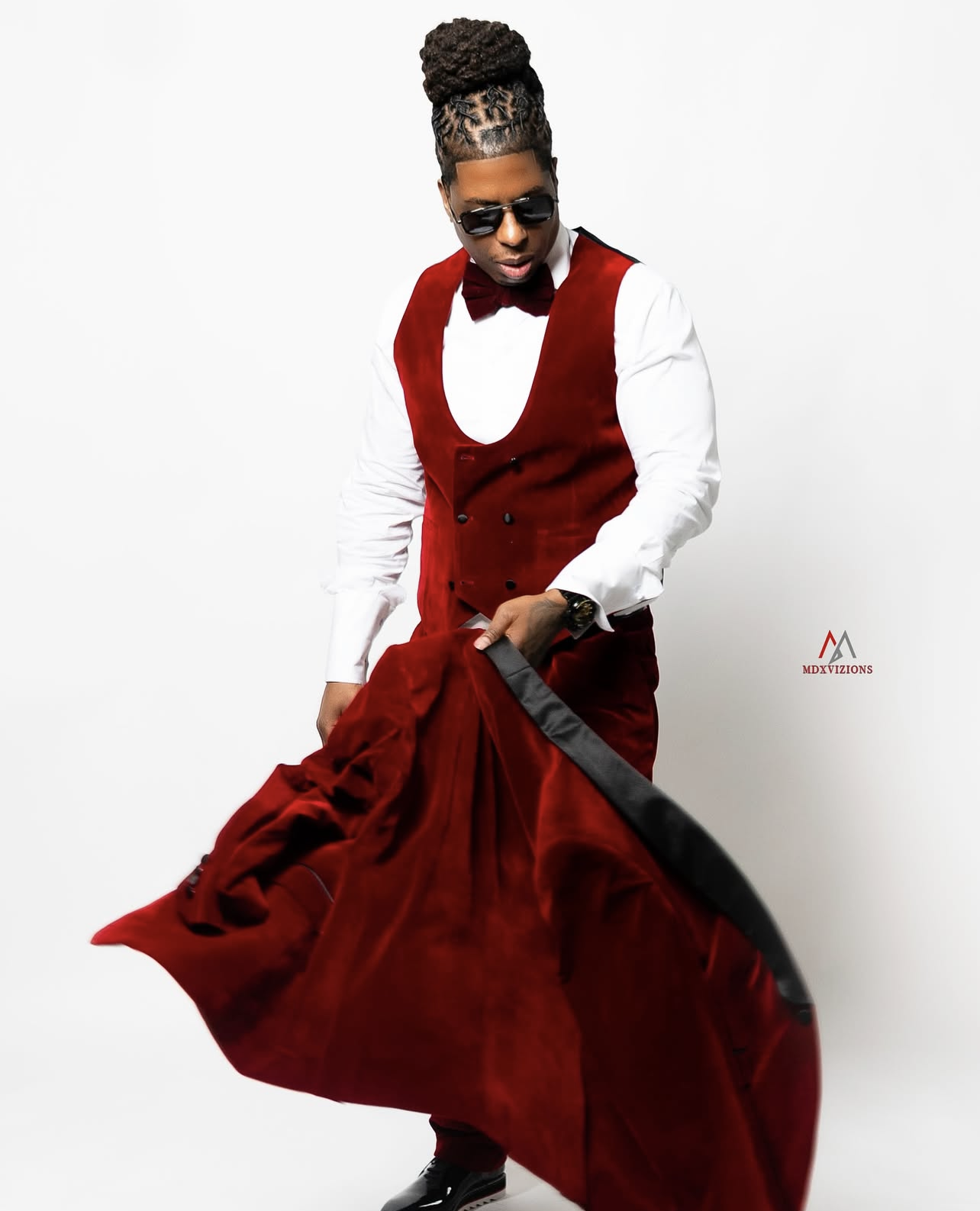 A man with dreadlocked hair styled in a high bun wearing sunglasses, a white shirt, a burgundy vest, and matching pants, holding the edge of his flowing red velvet coat.