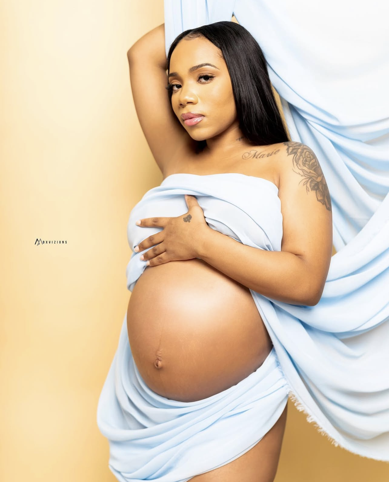 Pregnant woman with long hair, tattoos, and light makeup posed against a yellow background, draped in light blue fabric.