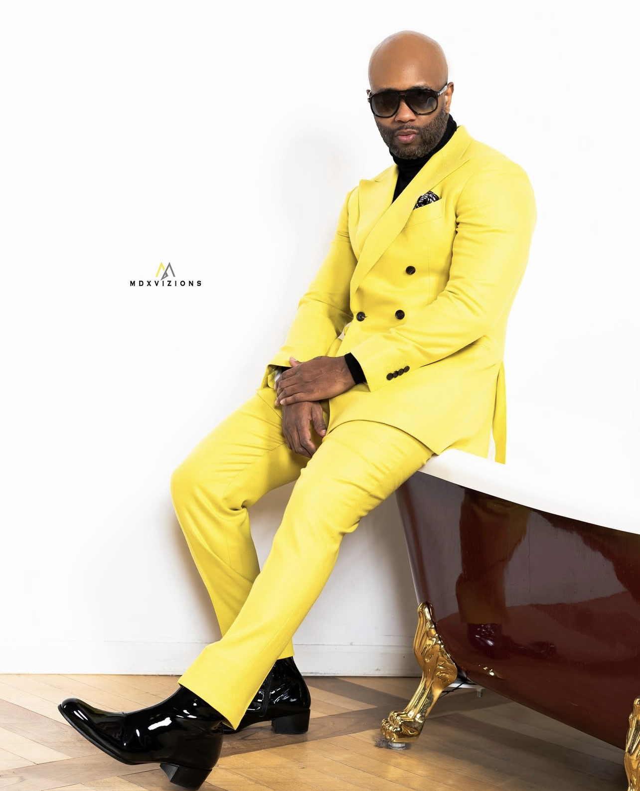 A man sitting on a white surface next to a clawfoot bathtub with gold details. He is wearing a bright yellow suit, black shirt, black sunglasses, and shiny black boots.