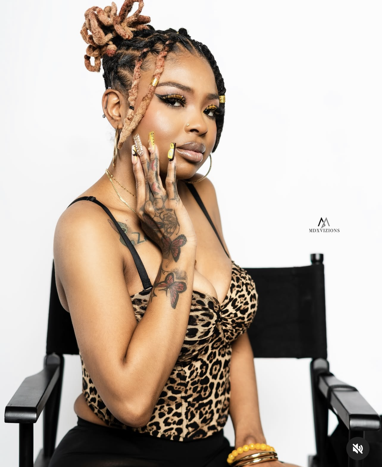 A woman with dreadlocked hair styled in a high bun, wearing a leopard print top, gold jewelry, and colorful nail polish, seated in front of a plain white background.