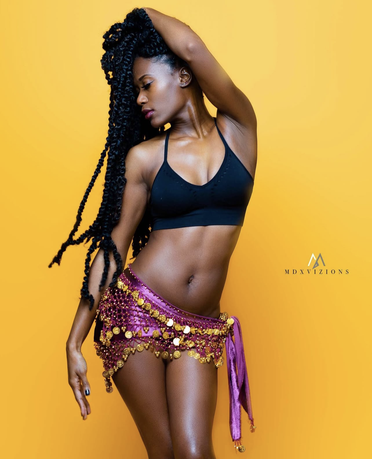 A woman with long, styled braids poses against a bright yellow background. She is wearing a black sports bra and a pink skirt decorated with gold coins and beads.