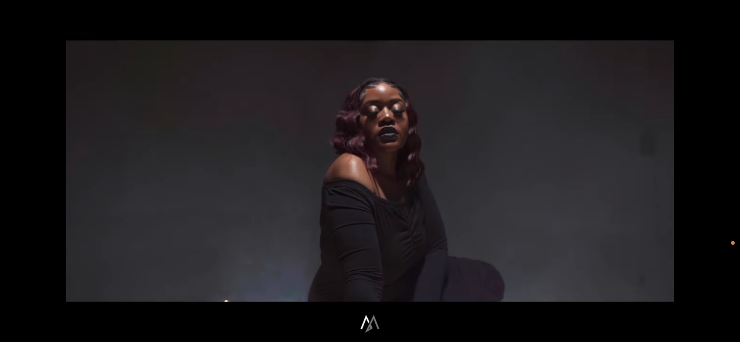 A woman with purple hair and dark lipstick sits against a dark and smoky background, wearing a black off-the-shoulder top.