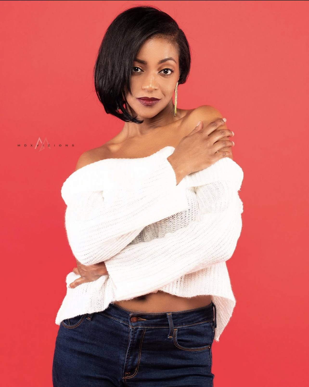 A woman with dark hair wearing a white off-the-shoulder sweater and dark jeans, standing against a red background.