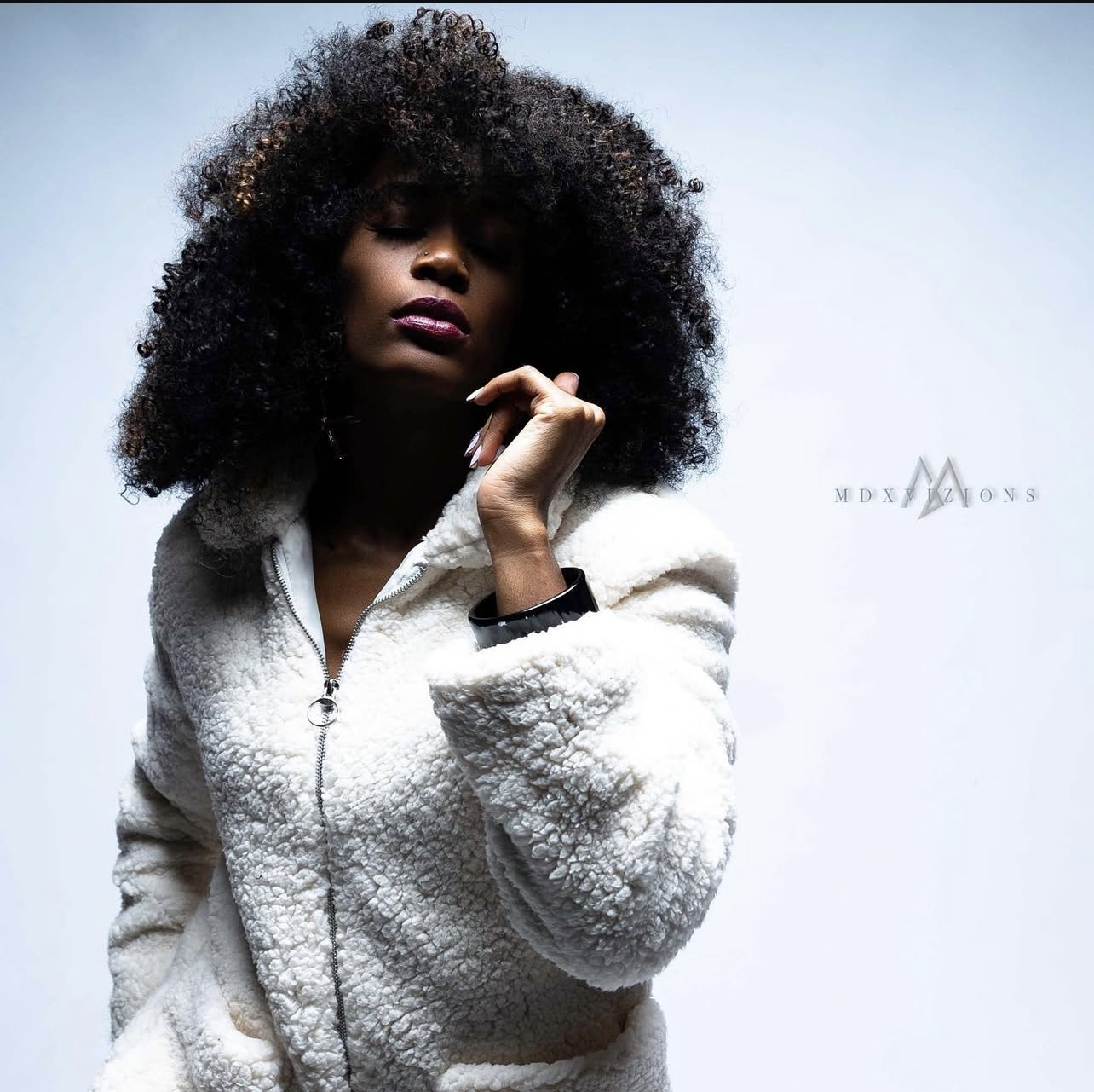 A woman with dark skin and curly hair wearing a white fuzzy zip-up jacket, posing with her eyes closed and her hand near her face against a plain background.