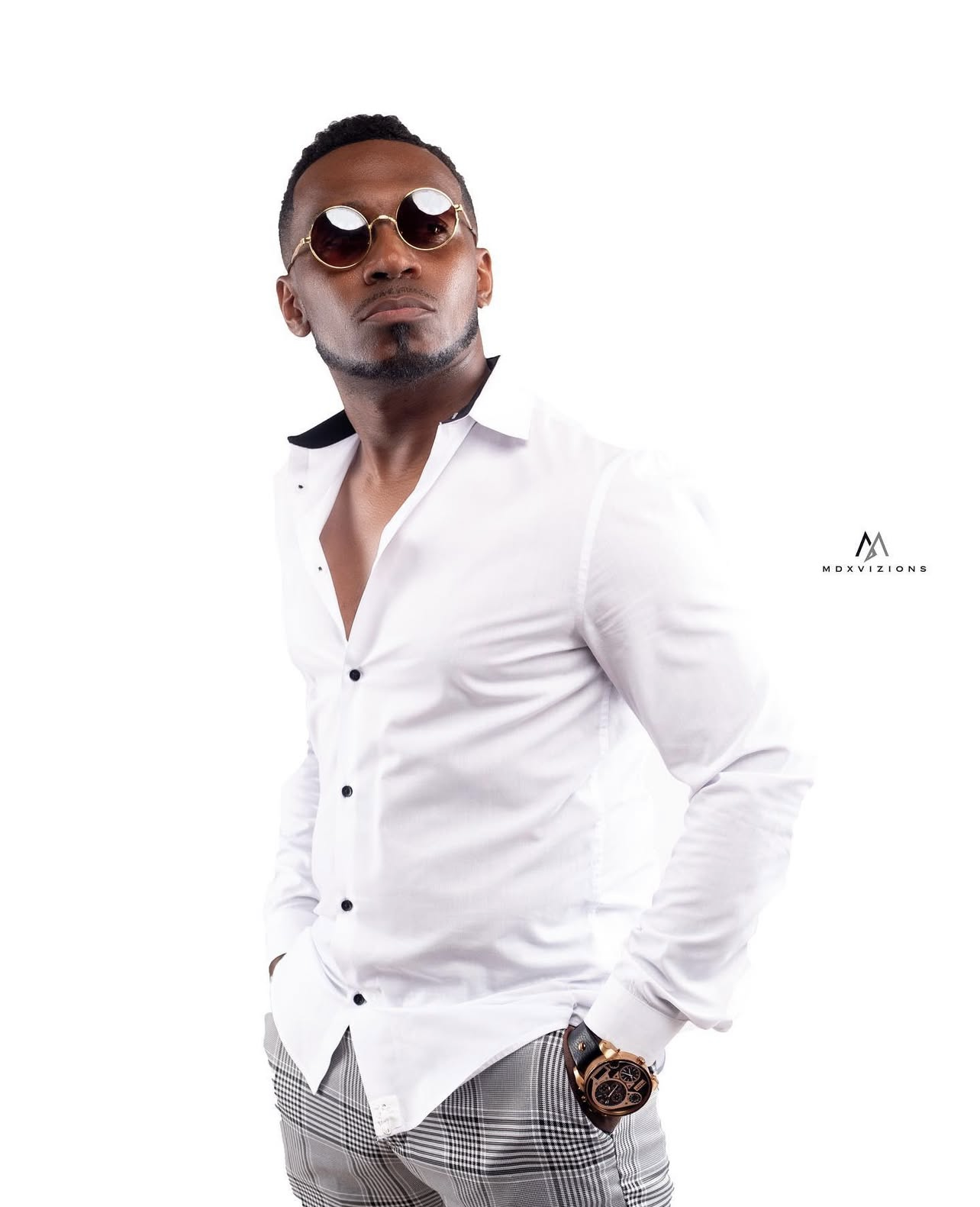 A stylish man wearing sunglasses, a white shirt with black buttons, patterned pants, and a watch, posing against a white background.