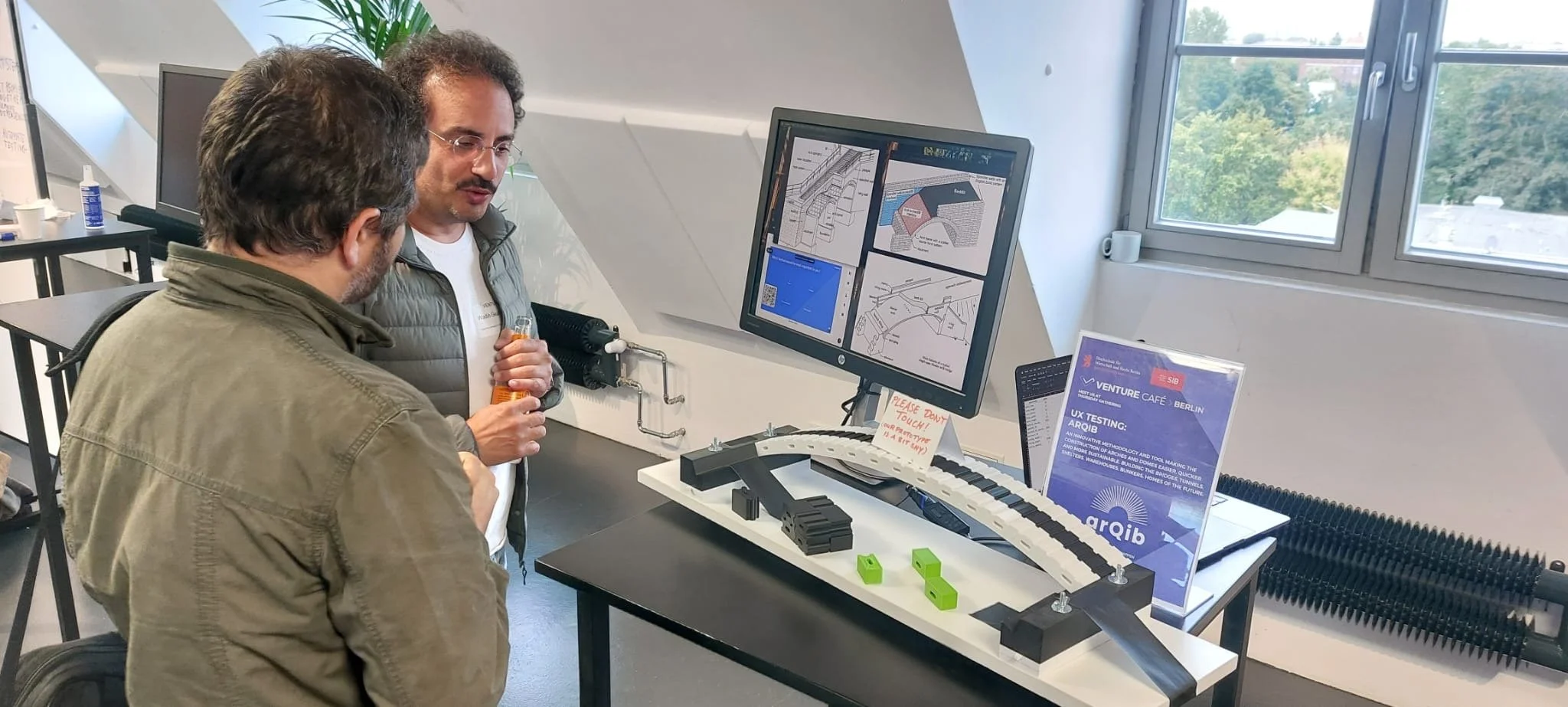 Two men are engaged in a discussion at a presentation table displaying a model of a curved architectural or engineering structure, with blueprints, signs, and a computer monitor showing technical drawings and plans.