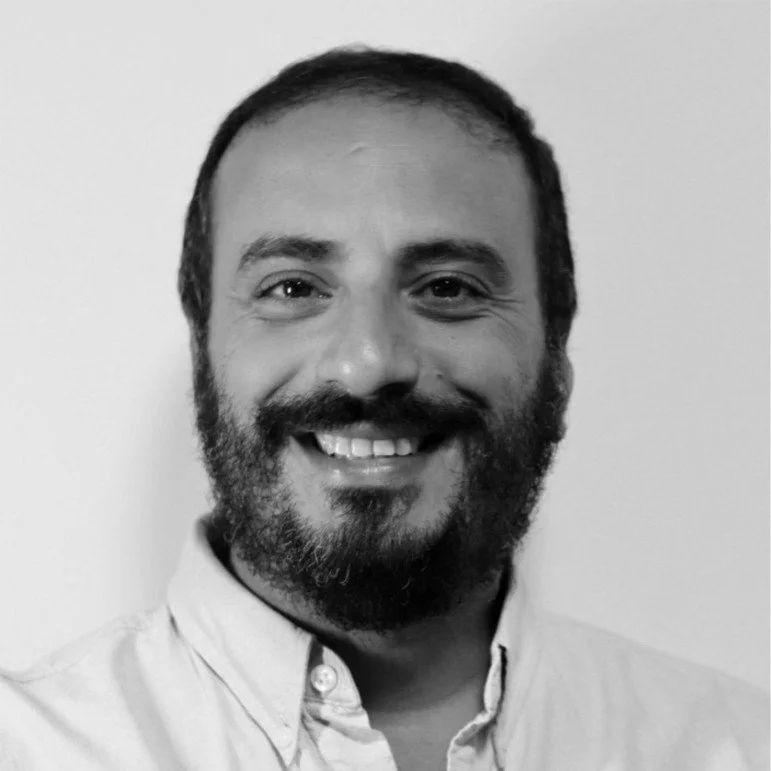 A smiling man with a beard and mustache, wearing a button-down shirt, standing in front of a plain light-colored background.