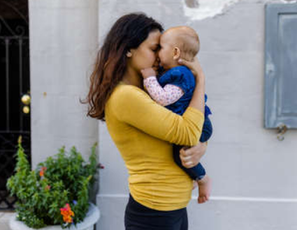 A woman in a yellow top holding and kissing a young child in a blue sweater outside near a gray wall and potted plants.