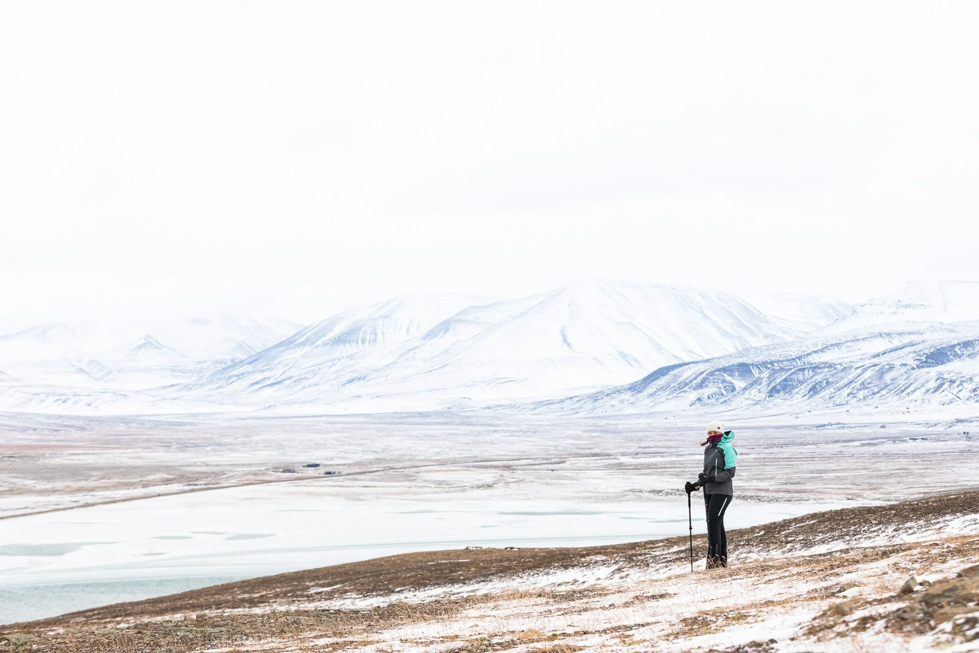 A person dressed in waterproof/windproof gear with a hat, holding walking poles, standing on a snowy landscape with mountains in the background, while the foreground is less snow-covered and brown colored with patches of snow.