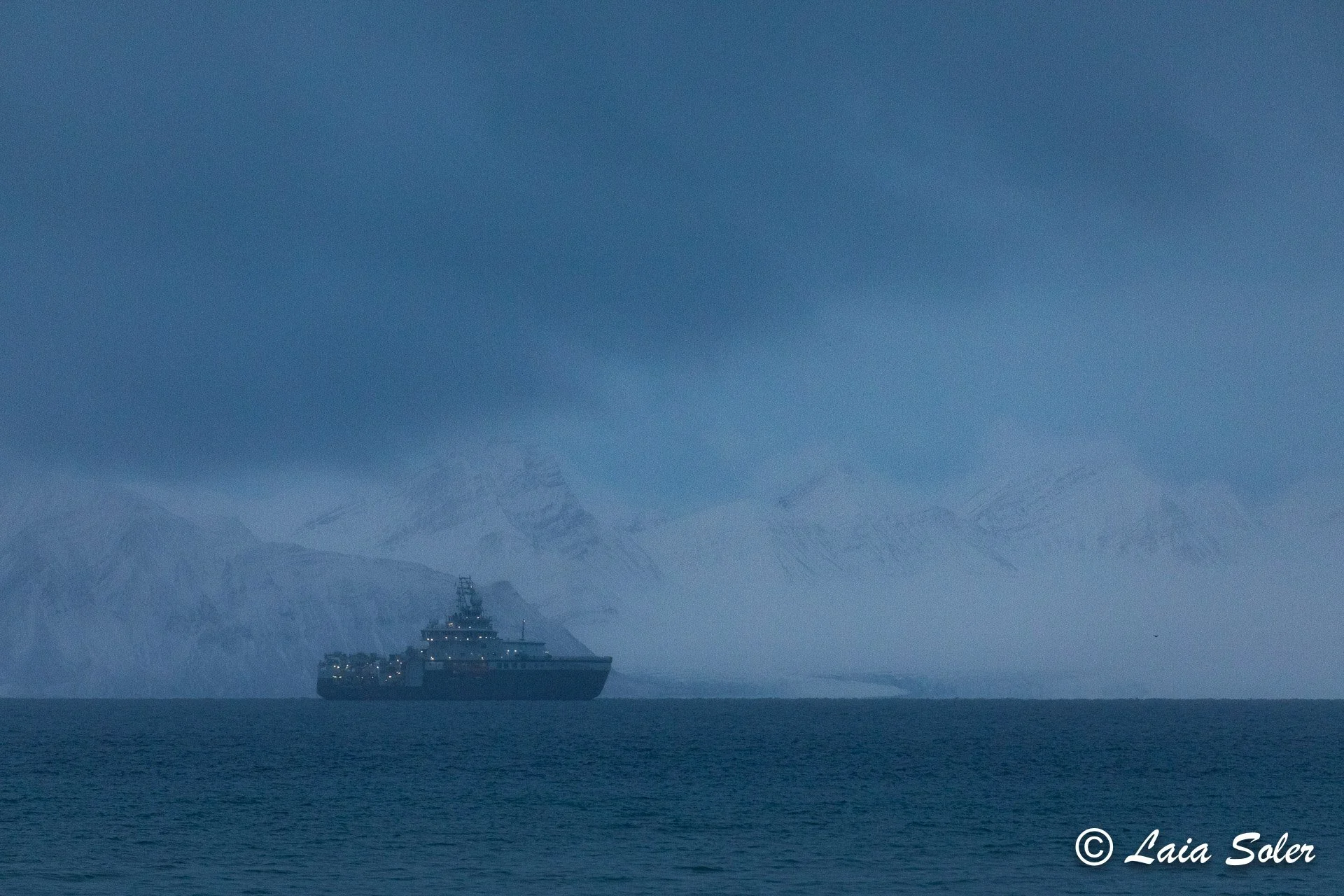 A large ship sailing in a vast ocean with snow-capped mountains in the background under a cloudy sky.