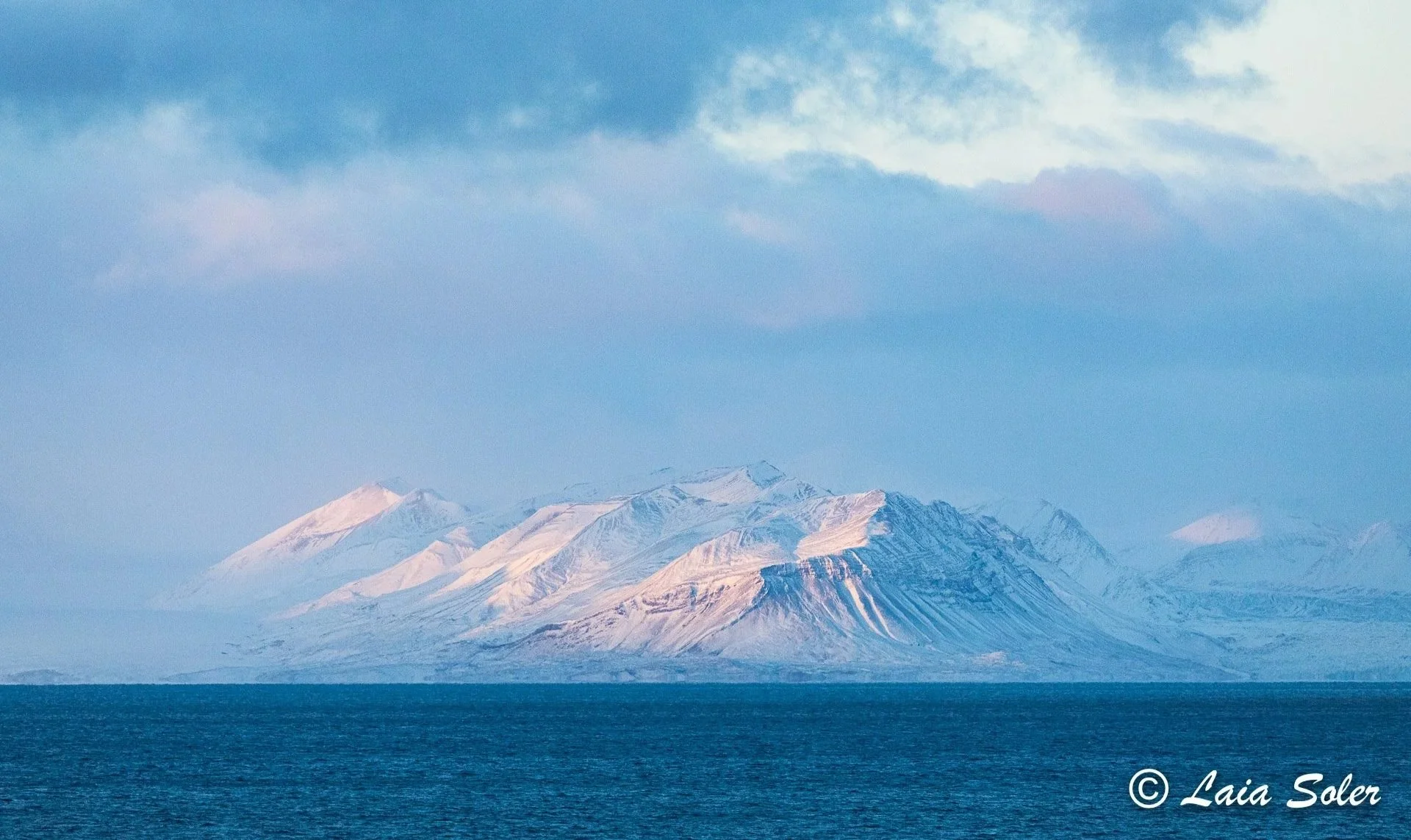 Snow-covered mountains across a body of water with clouds in the sky.