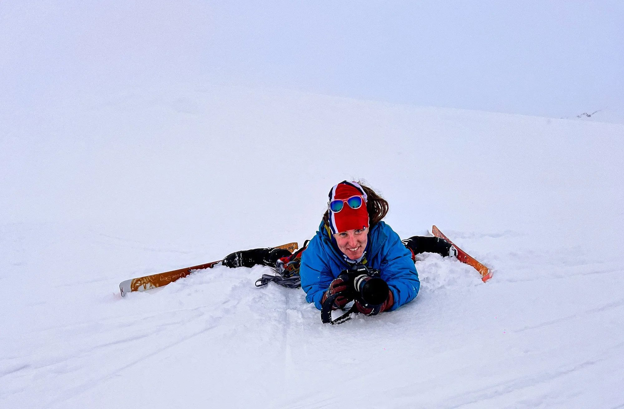A person lying in the snow with a camera, surrounded by skis, wearing a blue jacket, red headgear with sunglasses, in a snowy landscape.