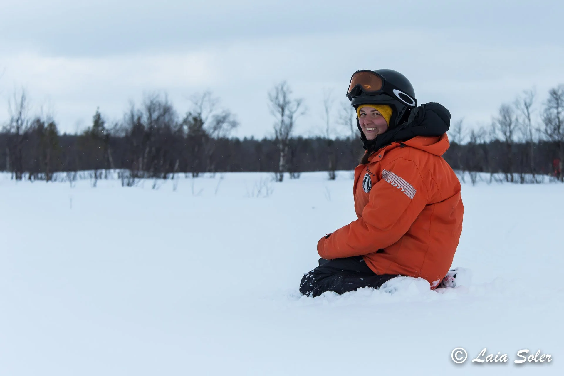 Person wearing an orange winter jacket with a black helmet and goggles, sitting on the snow in a winter landscape with bare trees and grey sky.
