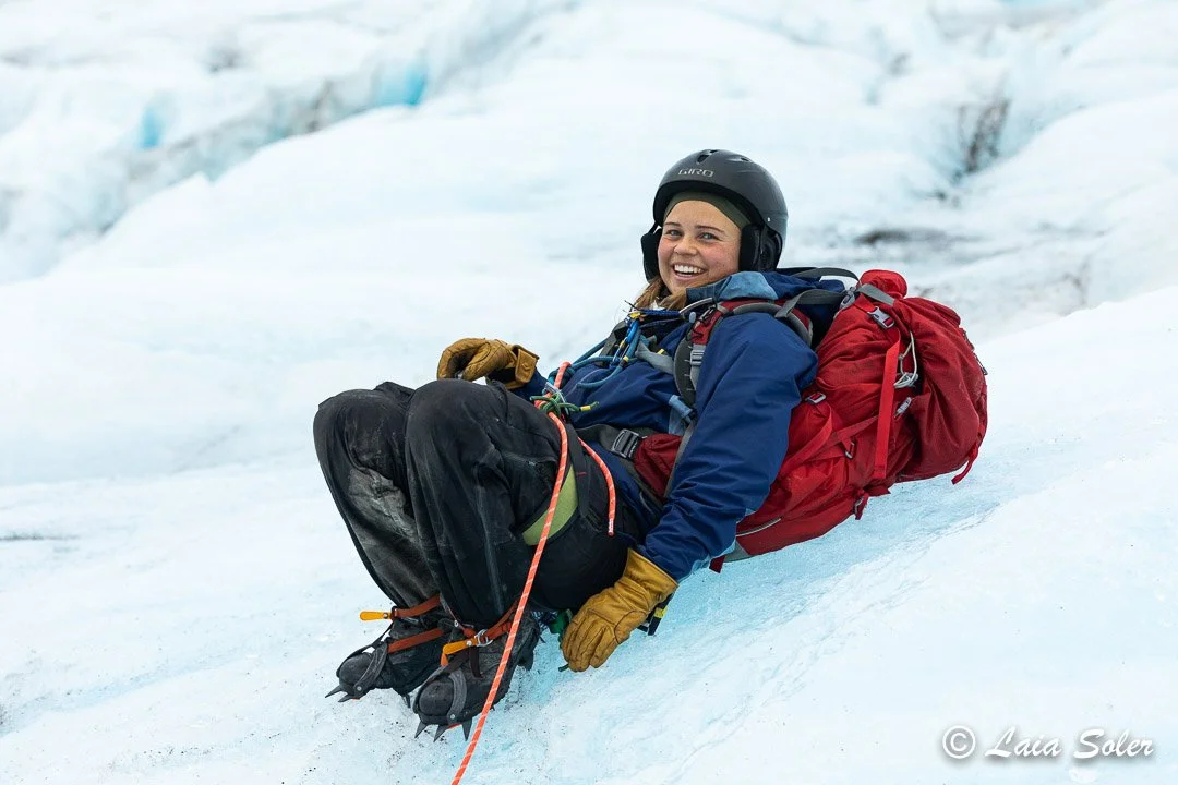 A female climber with a helmet, gloves, crampons and a red backpack resting on the glacier's icy slope, smiling at the camera.