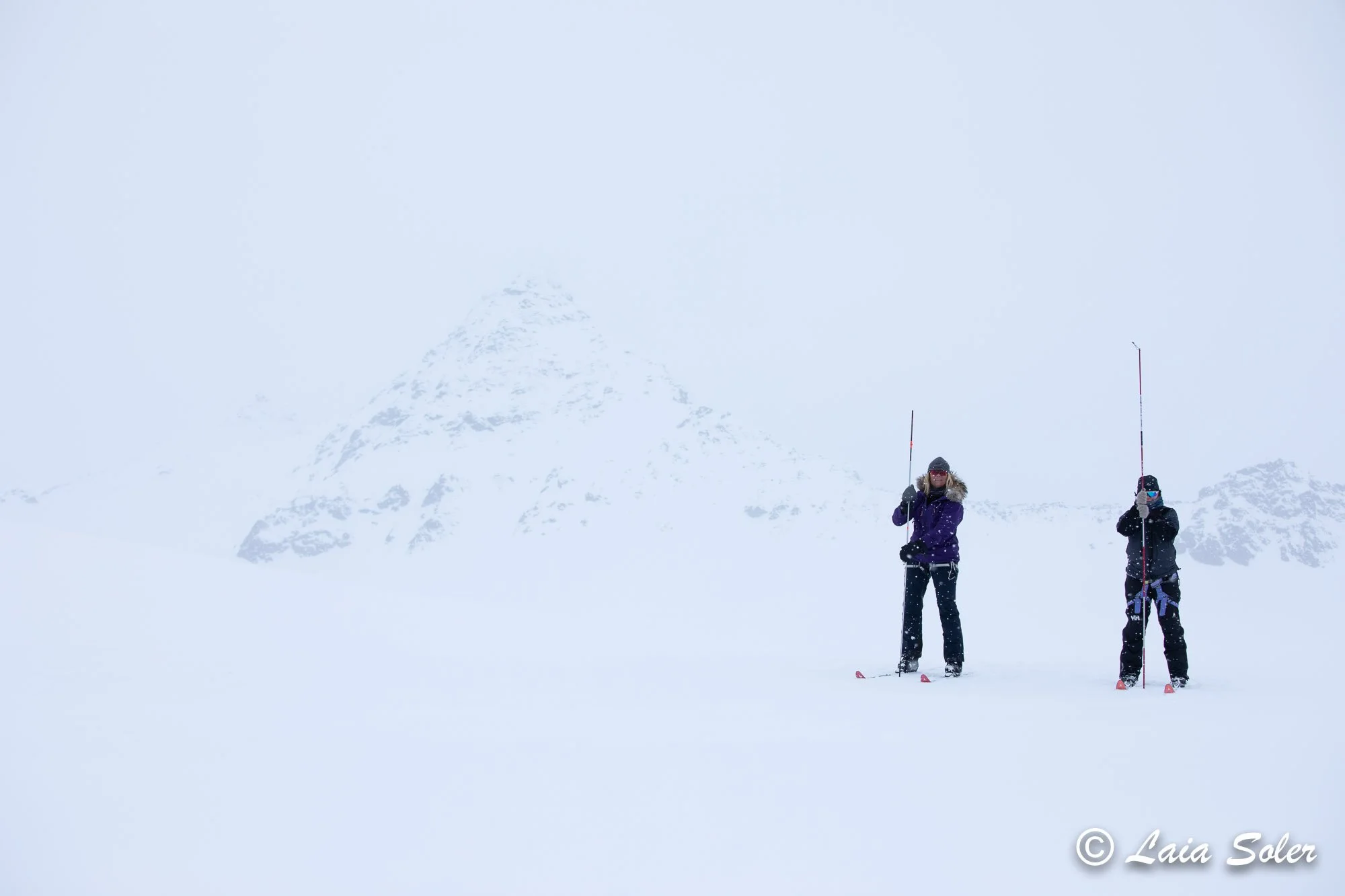 Two people dressed in winter clothing are standing on Nordic skis on a snowy landscape, with a snow-covered mountain in the background. They have probes to check for crevasses on the glacier they are standing on during a lunch break.