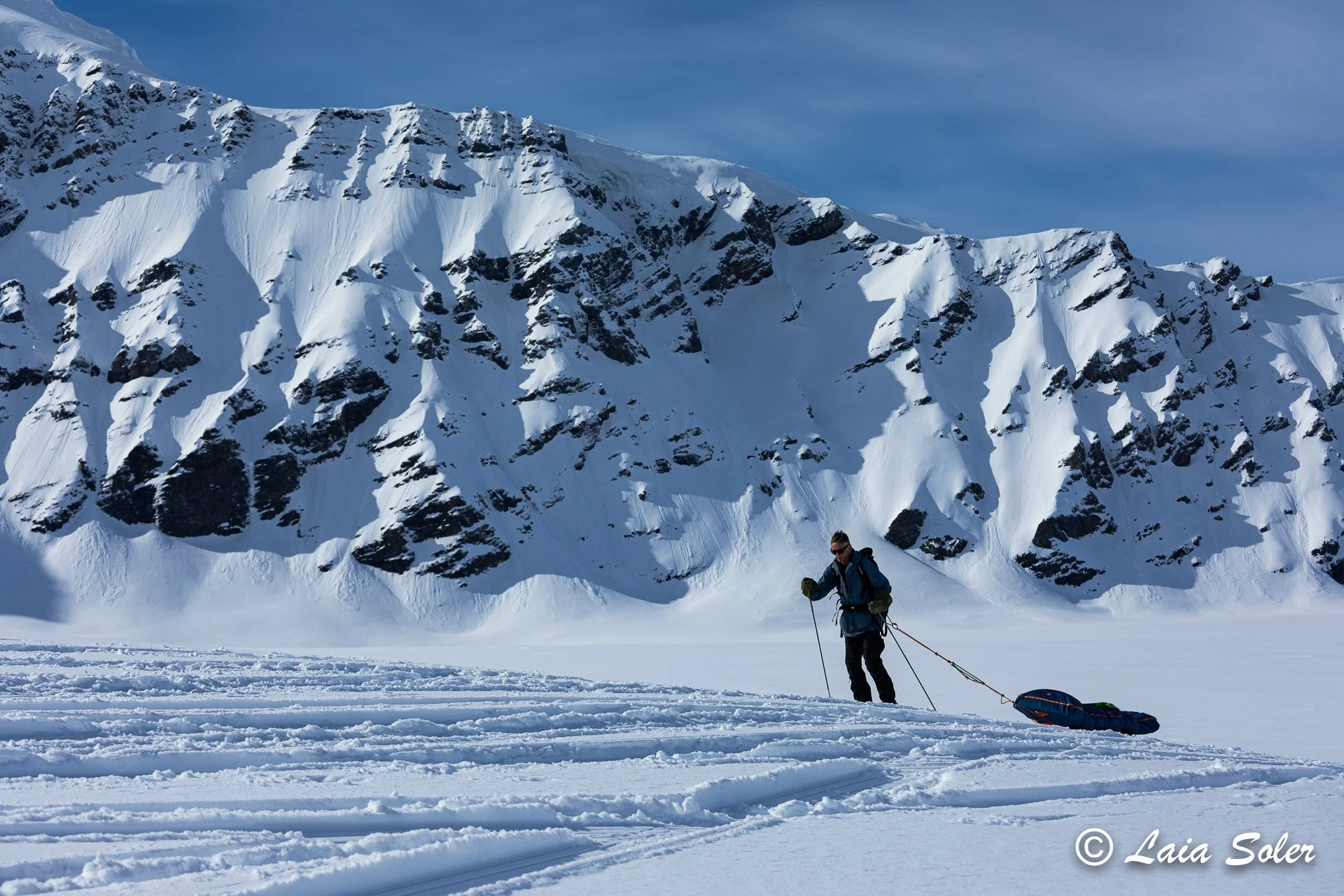 A person in winter gear and sunglasses pulls a sled across snow in front of snow-covered mountains under a blue sky.
