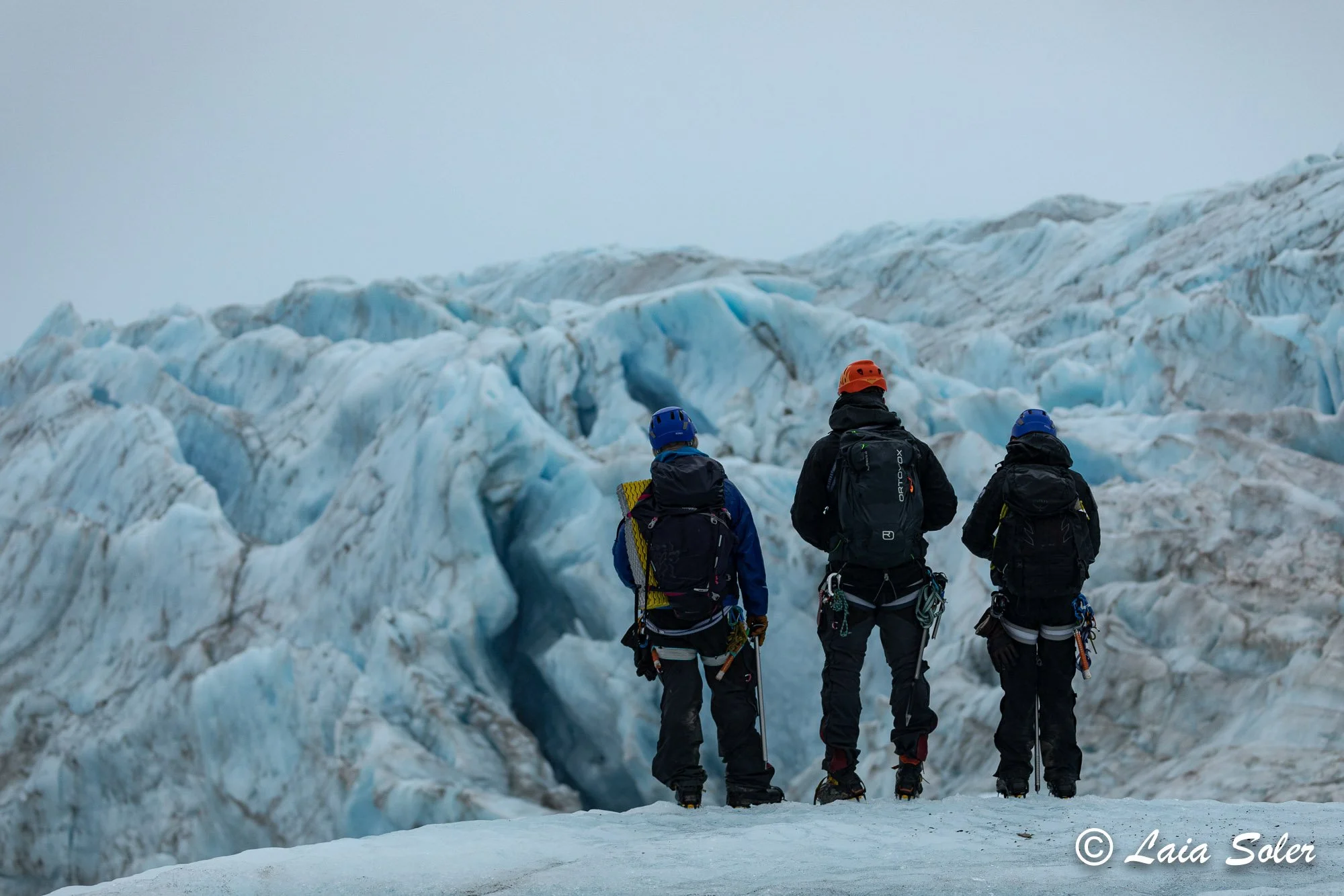Three mountaineers in winter gear stand on snow-covered terrain, looking at a glacier with jagged ice formations.