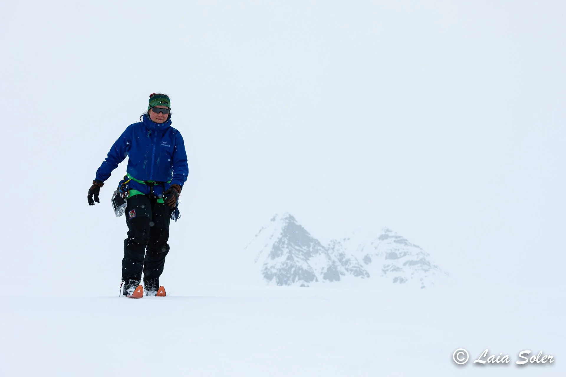 A person dressed in winter gear, including a blue jacket, black pants, and a green beanie, stands on skis on a snow-covered glacier with mountains in the background.