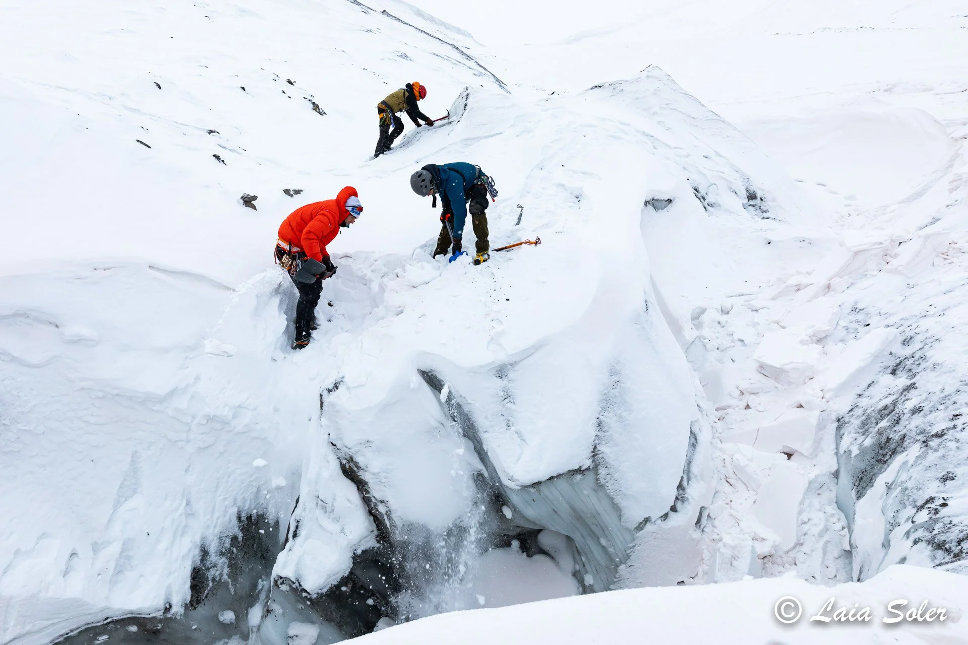 Three people dressed in winter clothing and wearing safety gear are climbing and working around a deep meltwater channel on a glacier.