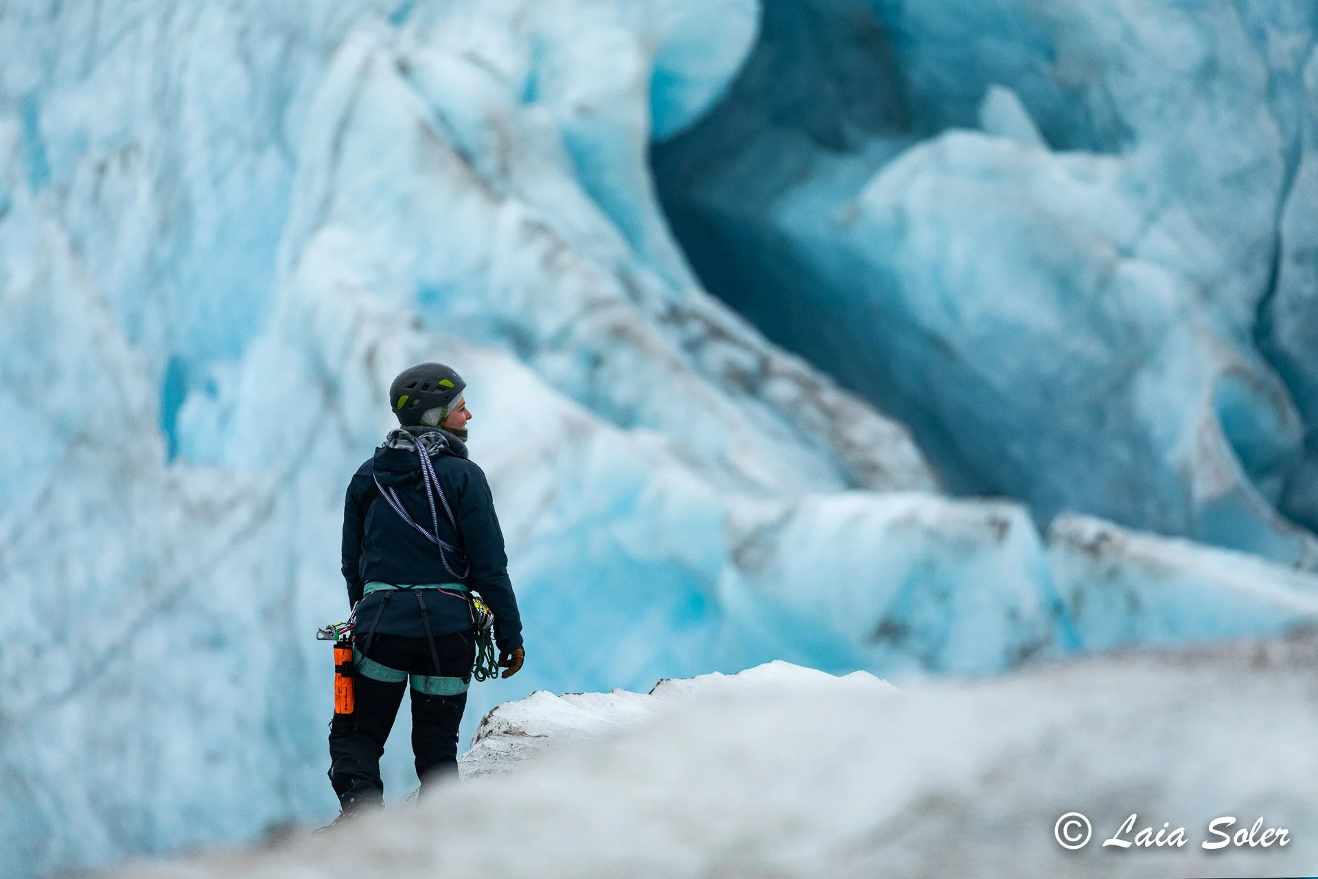 A person dressed in winter gear is standing in front of blue and white glacier ice, looking up at the icy formations.